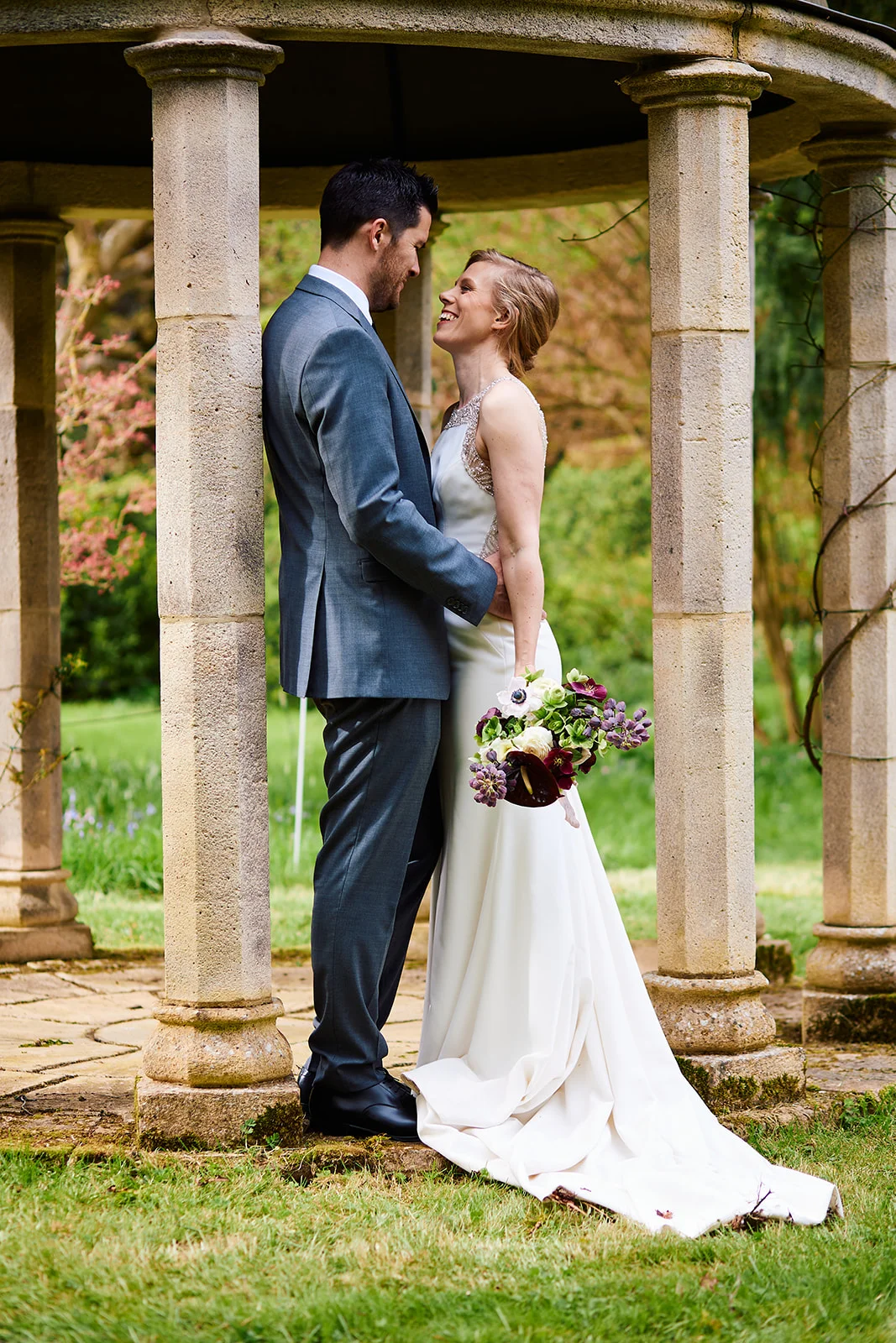 A newlywed couple laughing together while standing inside a stone gazebo holding a bouquet.