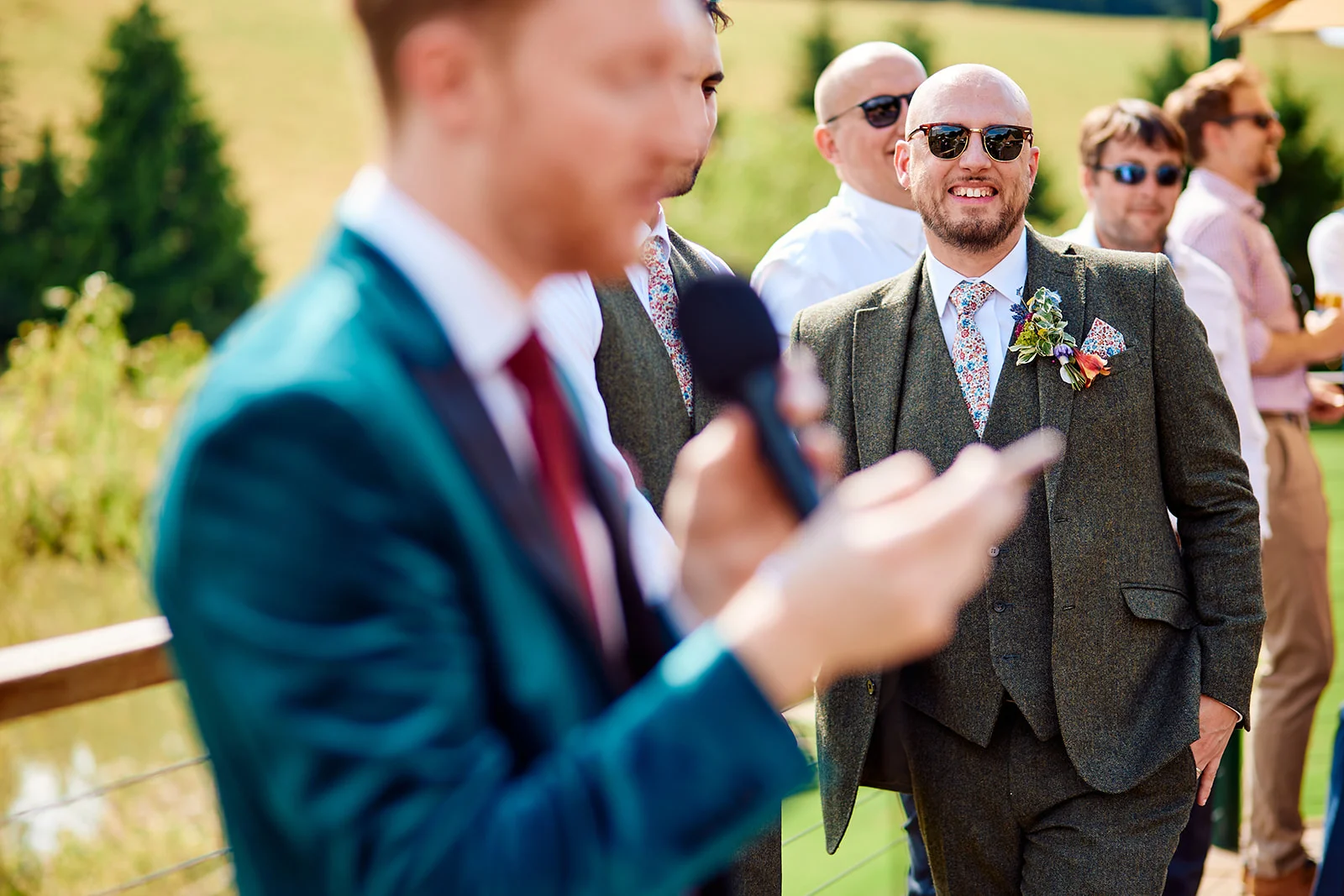 A groom in a tweed suit and floral tie smiles and laughs while listening to a wedding speech outdoors on a sunny day.