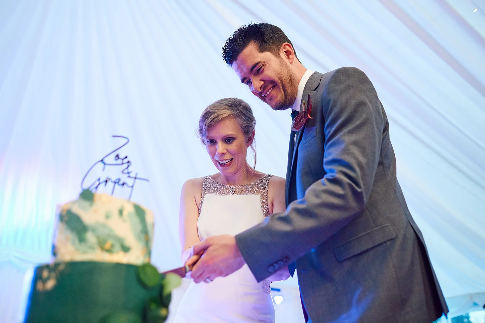 A smiling bride and groom cut into a decorative green wedding cake together inside a white marquee.