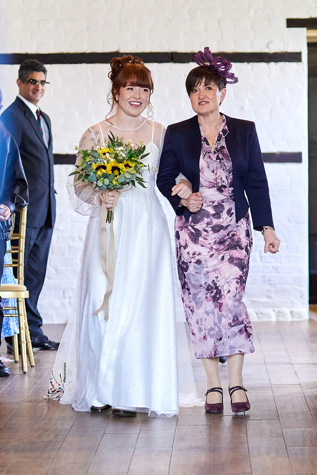 A smiling bride holding a sunflower bouquet walks down the aisle arm in arm with her mother, who wears a floral dress and fascinator.