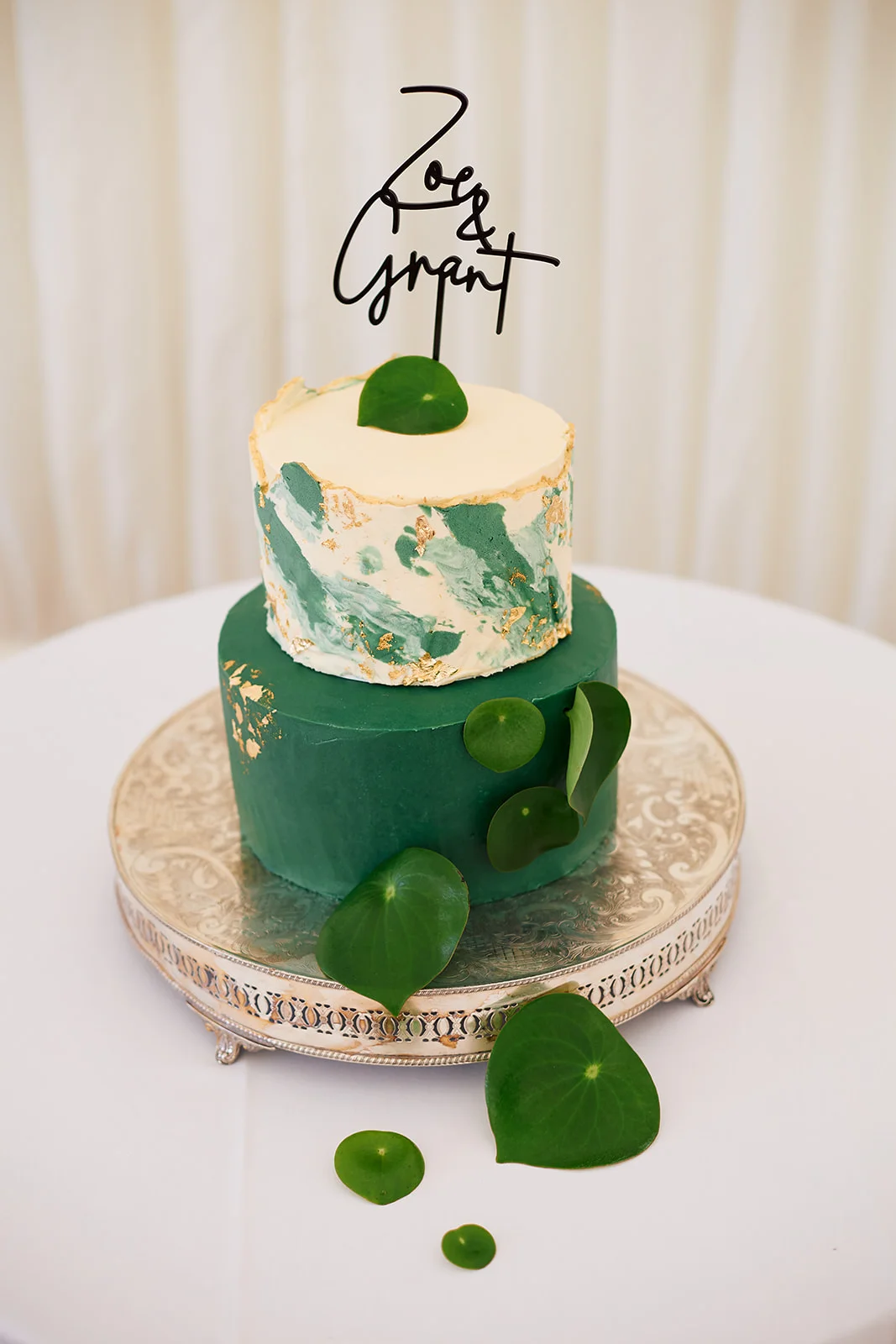 A stunning two-tier wedding cake featuring emerald green and marble-effect buttercream tiers decorated with gold leaf and fresh green leaves, topped with a modern black wire name topper on a silver ornate stand.