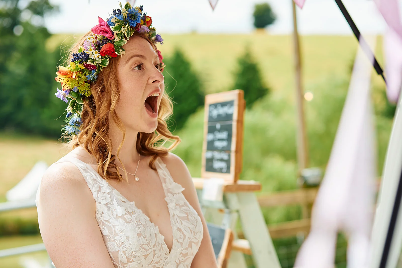 A joyful red-headed bride wearing a vibrant floral crown and a lace V-neck dress laughs excitedly during an outdoor festival wedding.