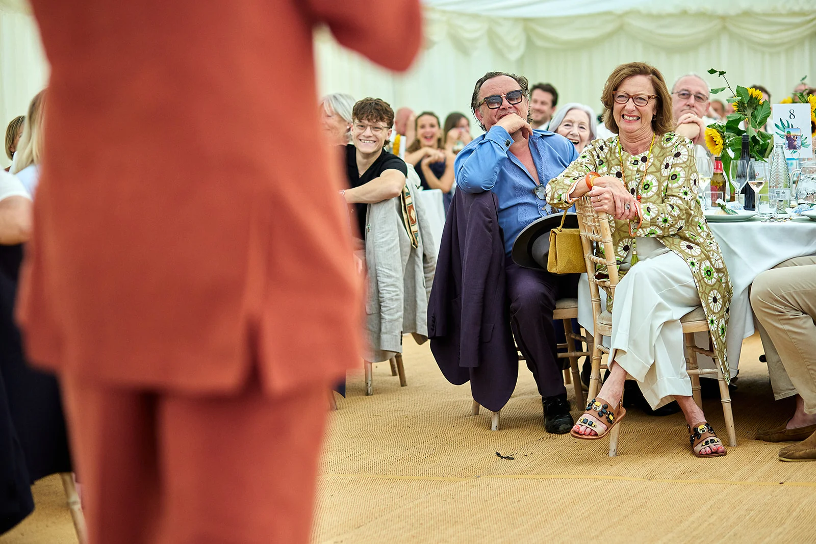 Wedding guests are captured laughing and enjoying speeches inside a bright marquee reception, viewed past the out-of-focus silhouette of a speaker.