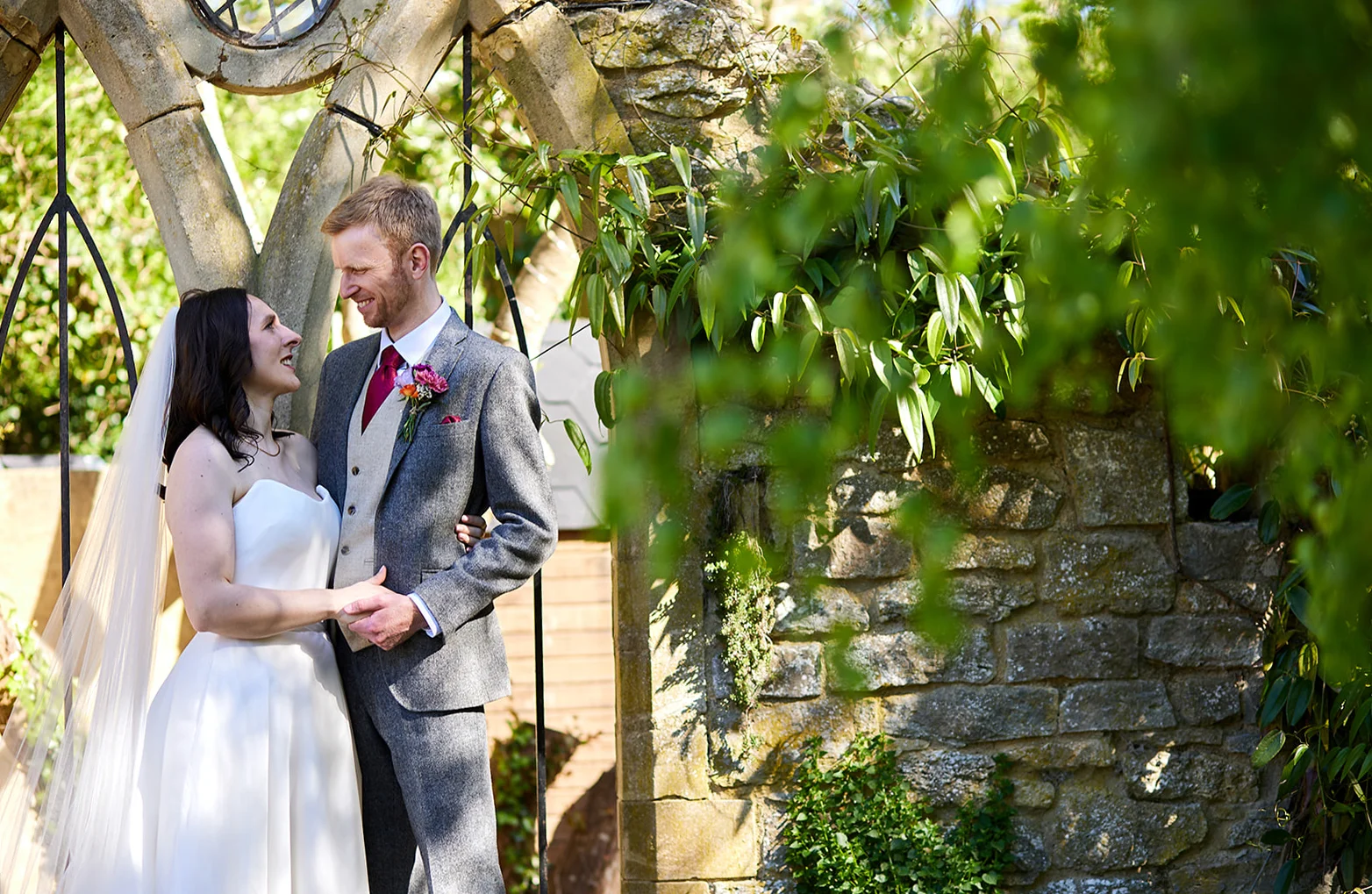 A smiling bride and groom share a tender look while standing together beneath an ancient stone archway framed by lush green foliage.