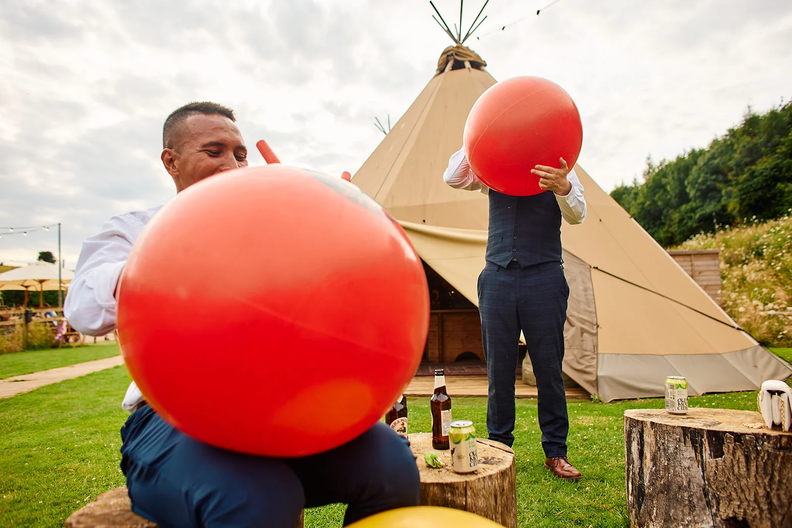 Groomsmen playfully inflate large red space hopper balloons outside a tipi wedding venue on a grassy lawn.