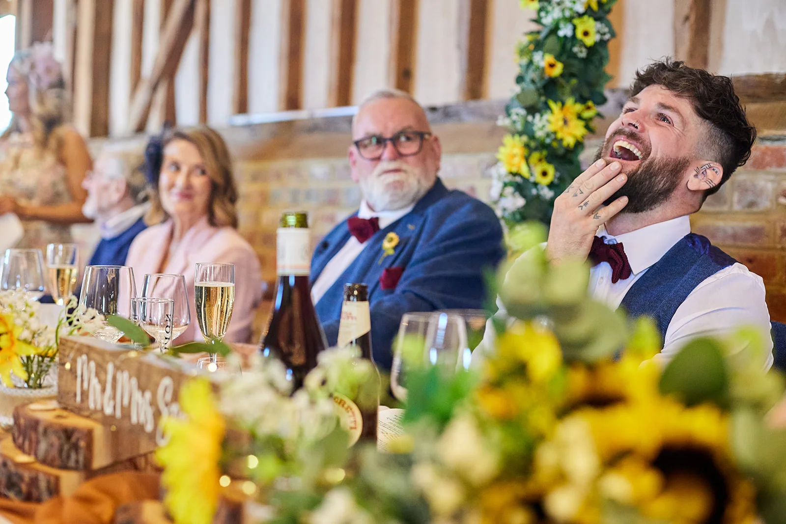 A groom with a beard and tattoos throws his head back in laughter during the wedding speeches at a rustic barn reception, with guests seated at the top table nearby.