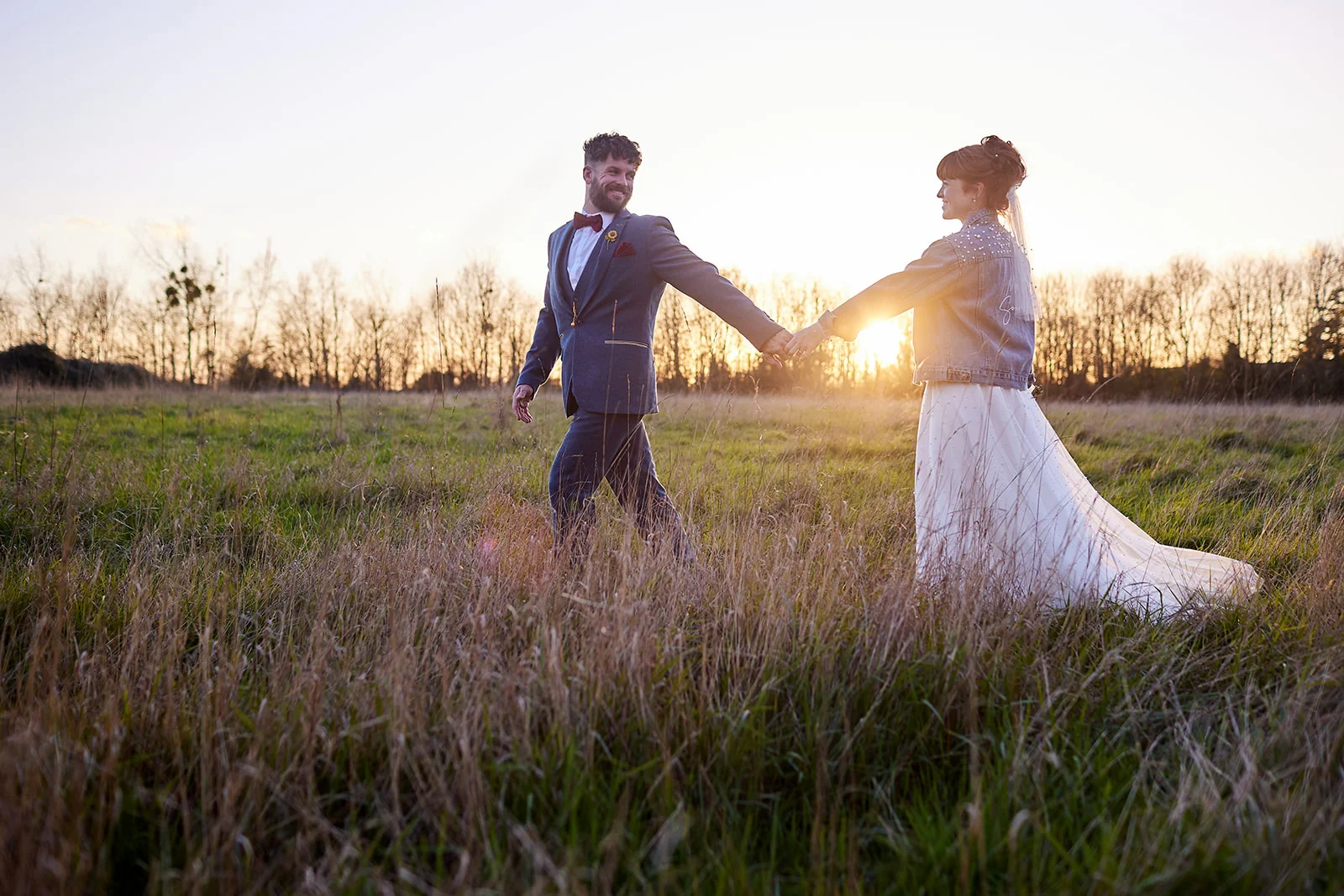A newlywed couple holds hands and shares a joyful glance while walking through a tall grass field during a glowing sunset.