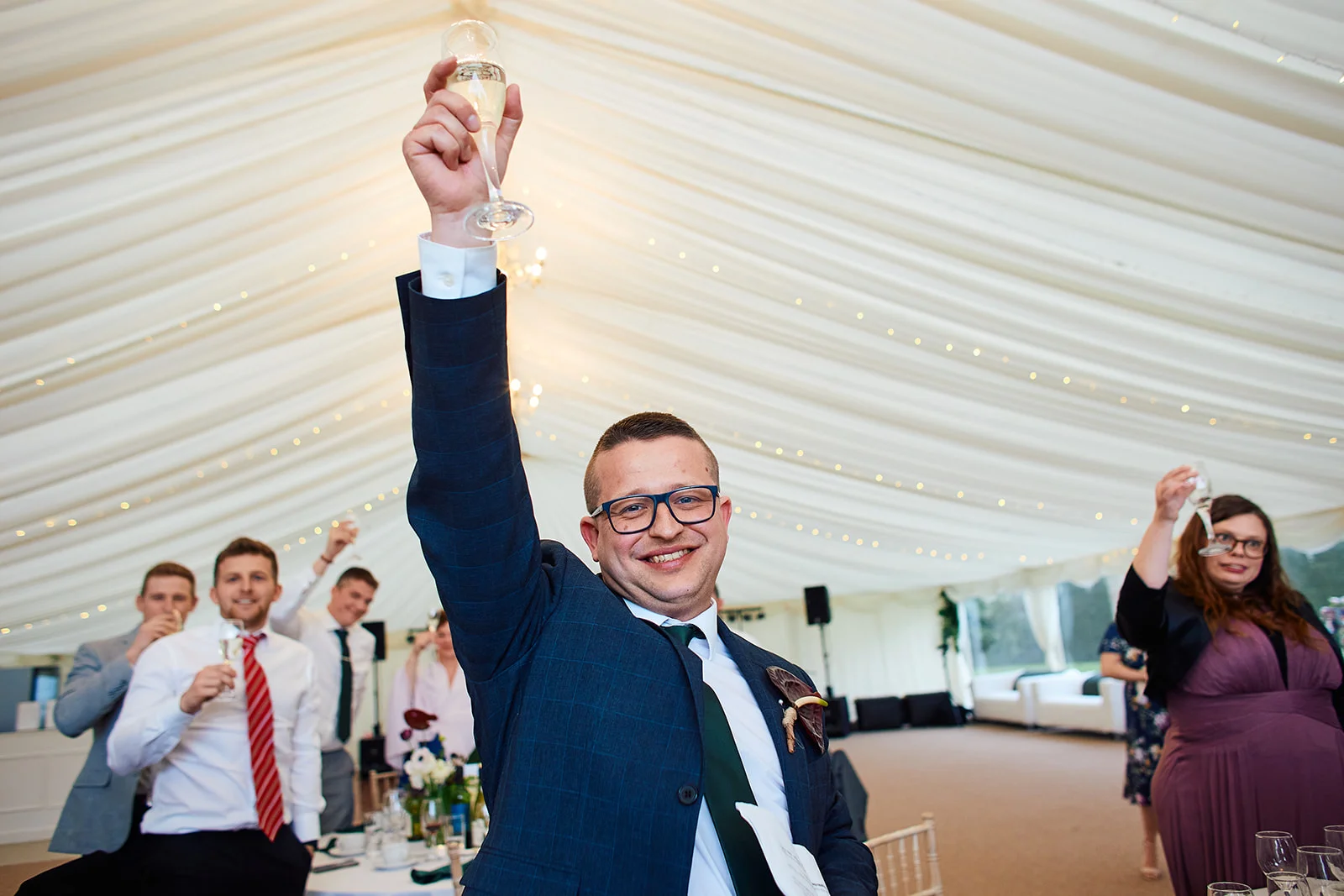 A smiling groom joyfully raises a glass of champagne during speeches inside a marquee draped with fabric and fairy lights.