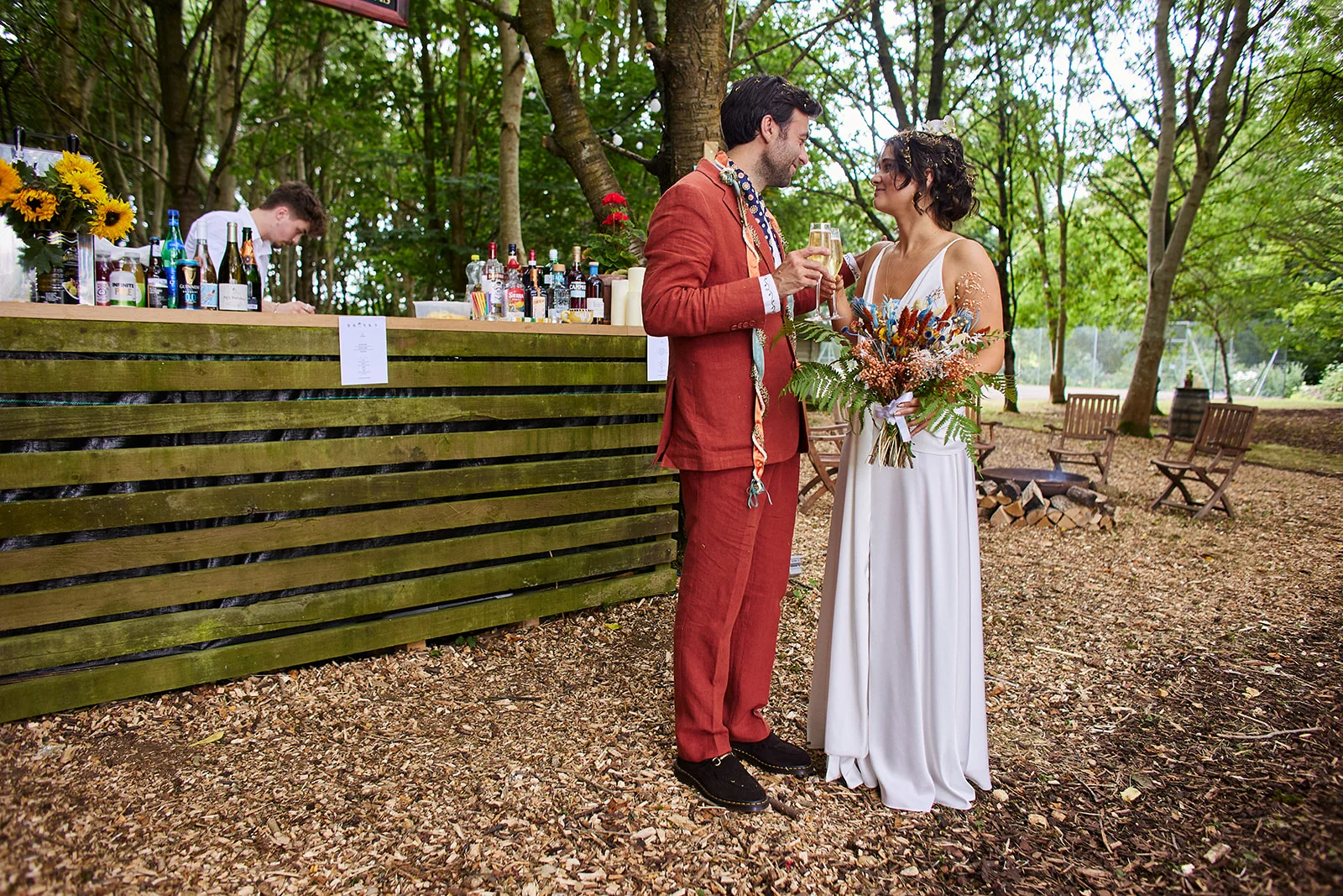 A bride in a white slip dress and a groom in a rust-coloured suit share a toast with champagne in a wooded outdoor setting near a wooden bar.
