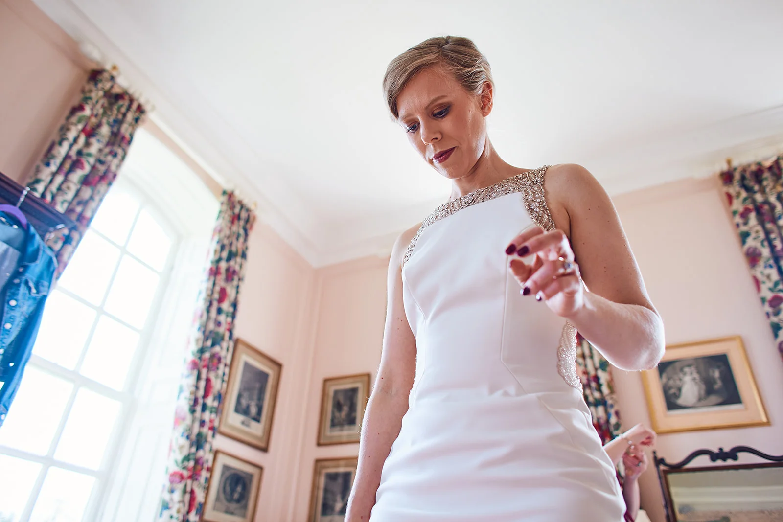 A low angle shot captures a bride with short hair looking down as she makes final adjustments to her crystal-embellished wedding dress during morning preparations.