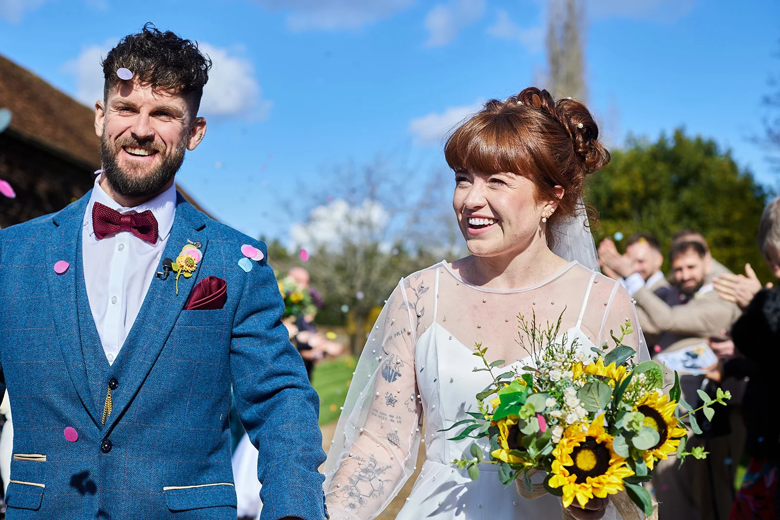 A joyful newlywed couple walks hand in hand through a shower of confetti on a bright, sunny day, with the smiling groom wearing a blue patterned suit and the bride holding a vibrant sunflower bouquet.