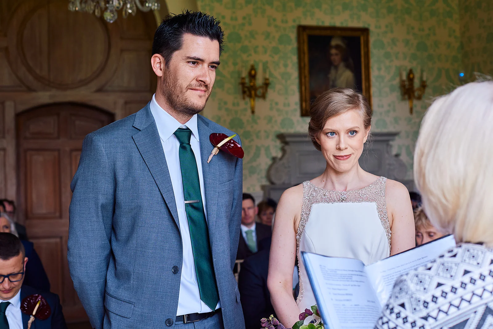 The bride and groom stand attentively during their civil ceremony inside an elegant wood-panelled room, with the groom wearing a grey suit and green tie.