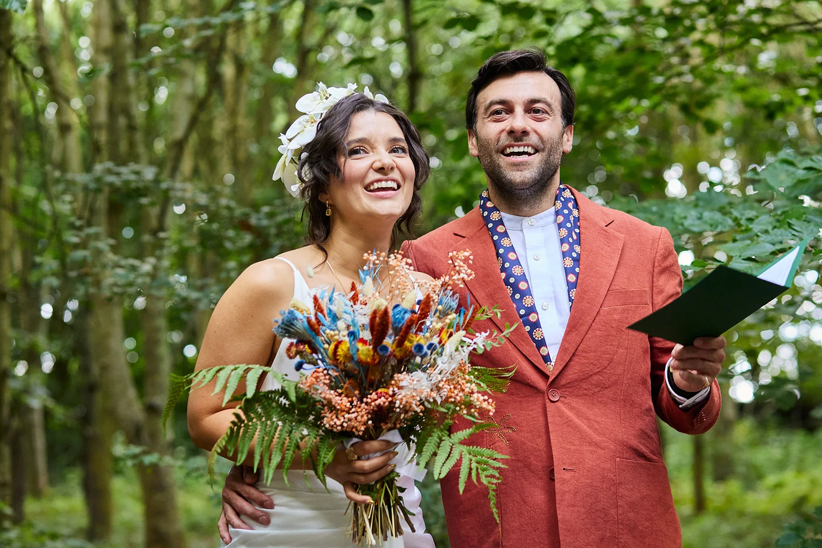 A joyful couple during an outdoor woodland ceremony, with the bride holding a colourful dried flower bouquet and wearing a floral crown, while the groom in an orange jacket reads from a vow book.