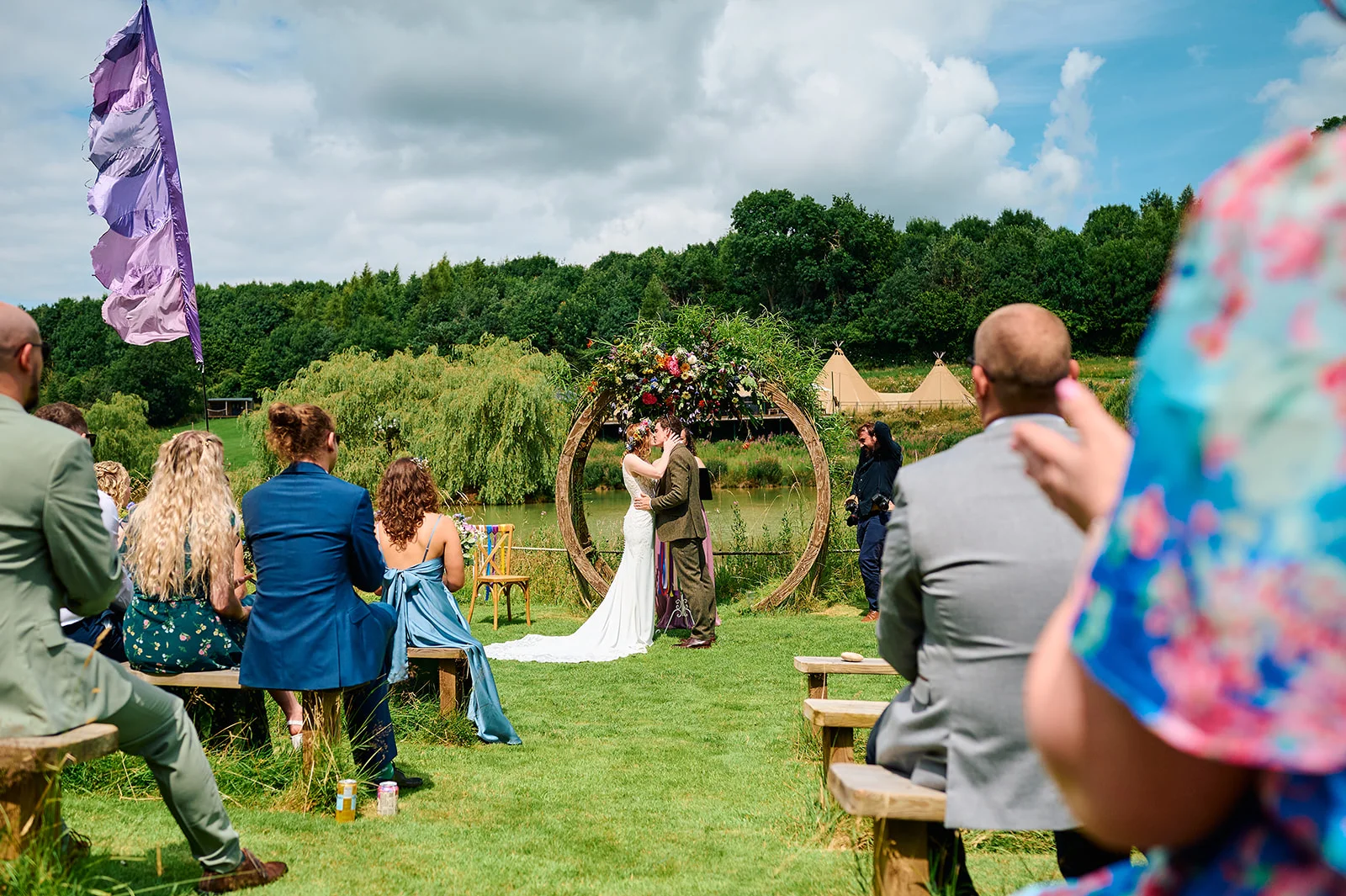 A bride and groom share their first kiss beneath a floral circular arch at an outdoor festival wedding by a lake.