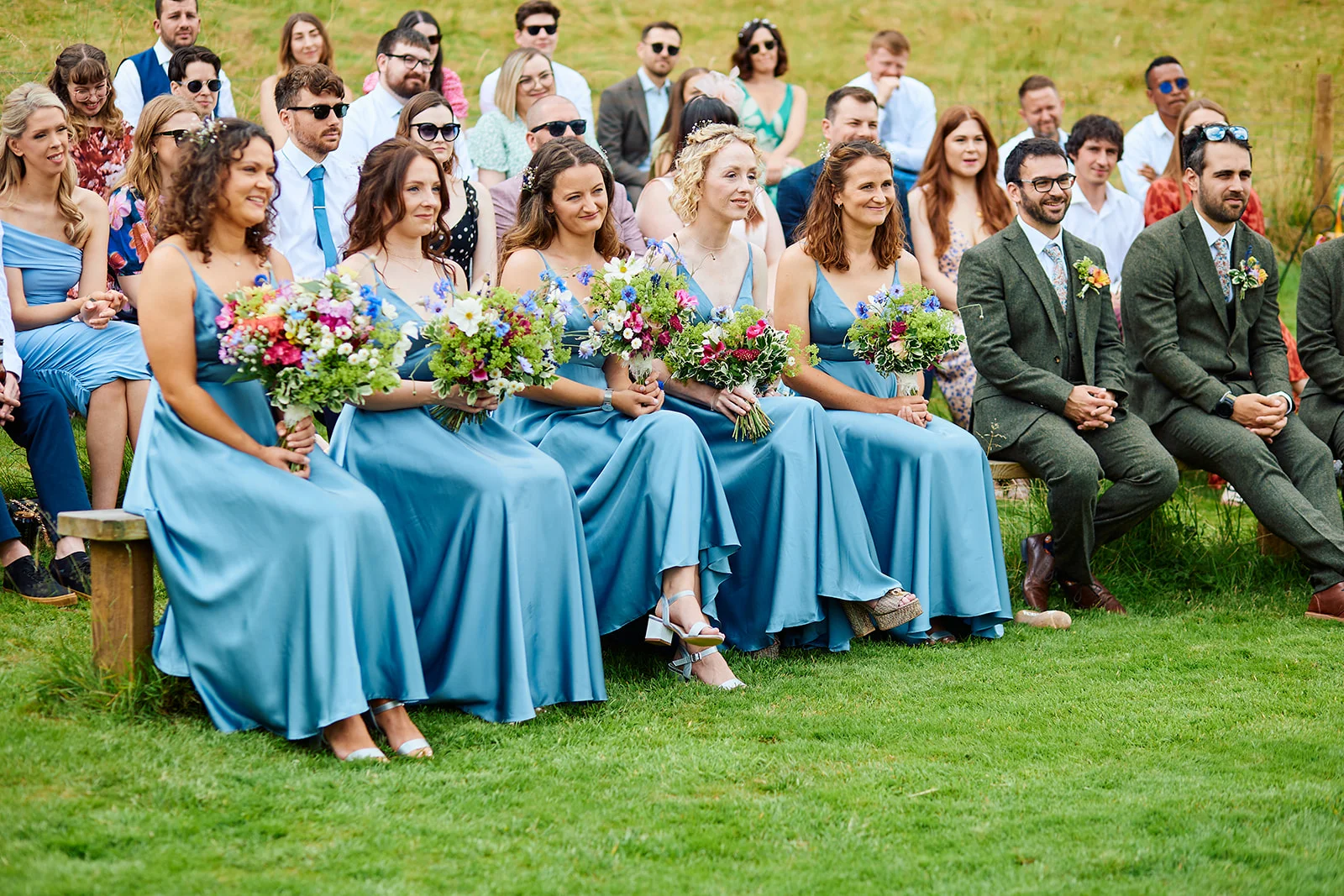 A bridal party of bridesmaids in dusty blue dresses and groomsmen in green tweed suits seated on benches during an outdoor wedding ceremony.