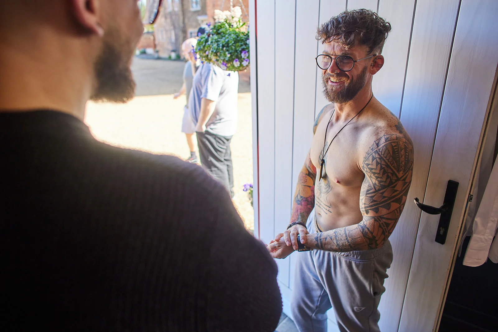 A tattooed groom smiles and laughs while getting ready indoors, viewed over the shoulder of another guest.