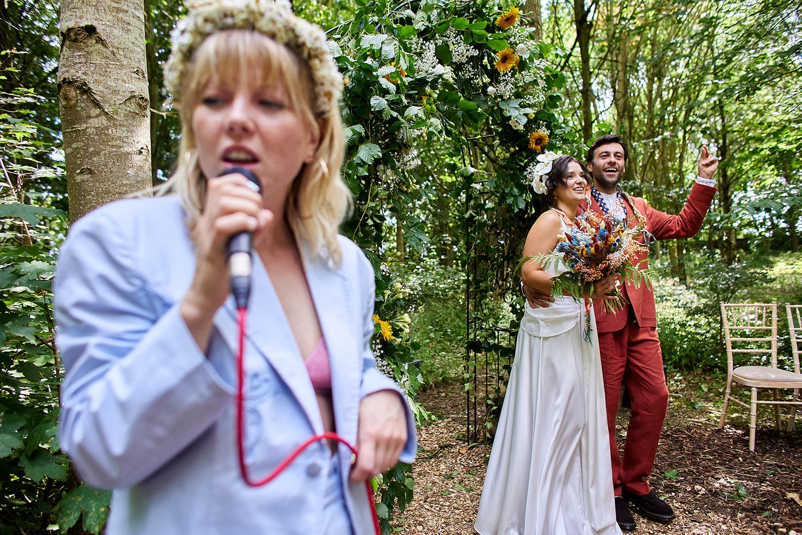 A live singer performs into a microphone in the foreground while a newly married couple celebrates under a floral arch in a lush woodland setting.