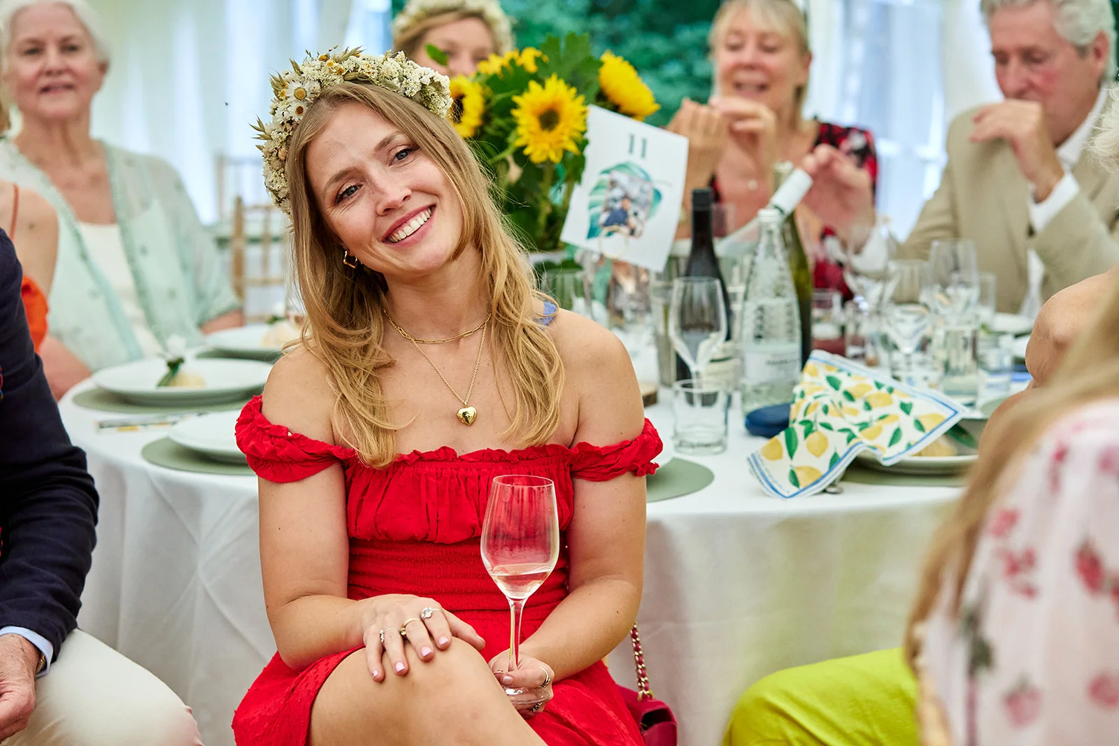 A smiling guest wearing a vibrant red off-the-shoulder dress and a floral crown holds a wine glass at a wedding reception table.