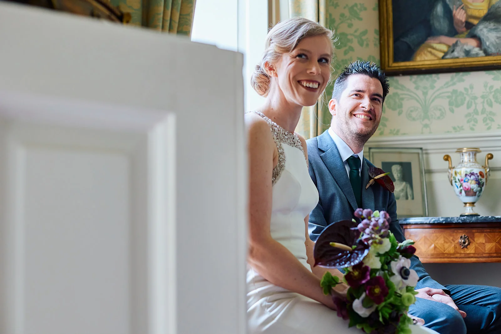 A smiling bride holding a dark floral bouquet sits next to her groom in a grey suit inside an elegant country house.