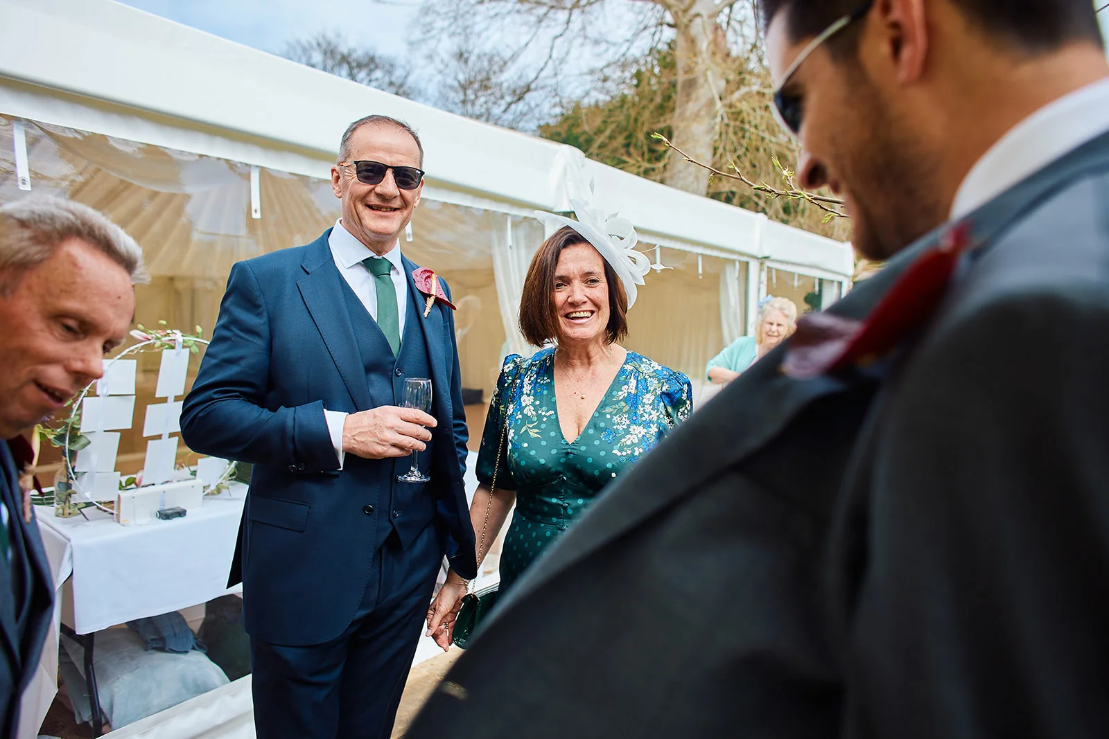 Smiling wedding guests and family members mingle outside a marquee during a drinks reception, holding drinks and laughing together.