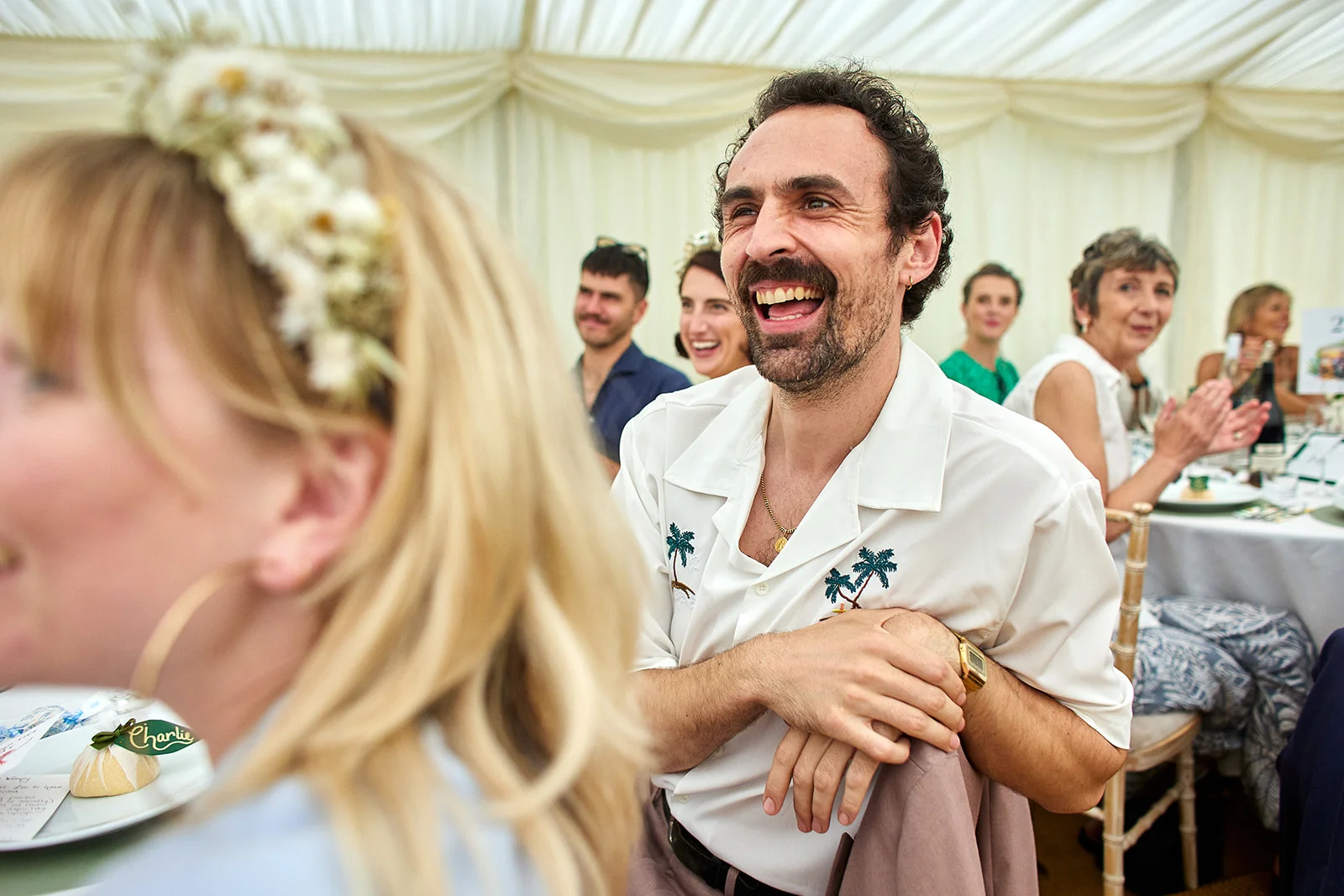 A smiling man with a moustache laughs joyfully while seated inside a draped marquee at a wedding reception.