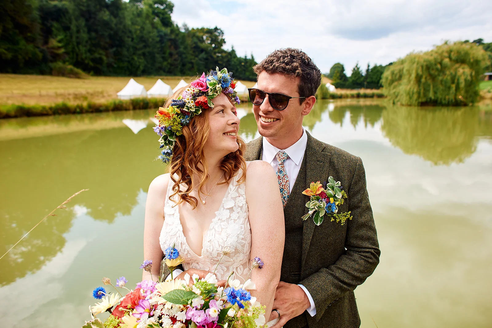 A newlywed couple shares a tender look by a scenic lake during an outdoor festival-style wedding.