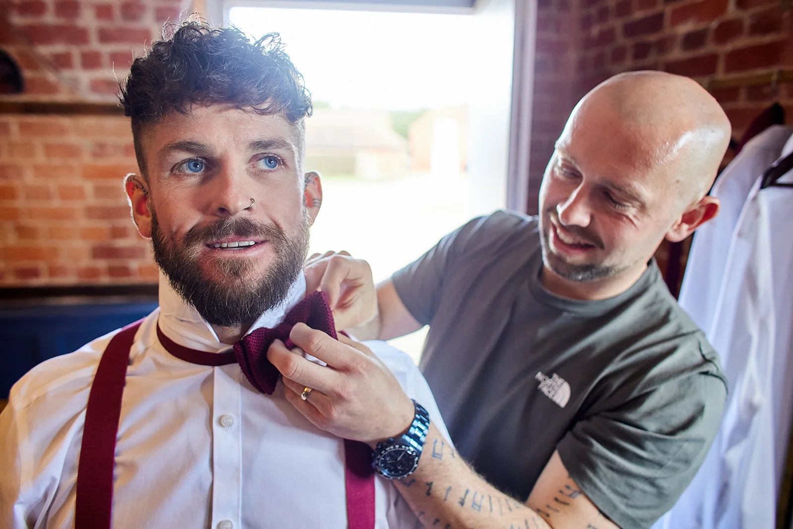 A groom with a beard and nose ring gets help adjusting his burgundy bow tie from a friend during wedding morning preparations.
