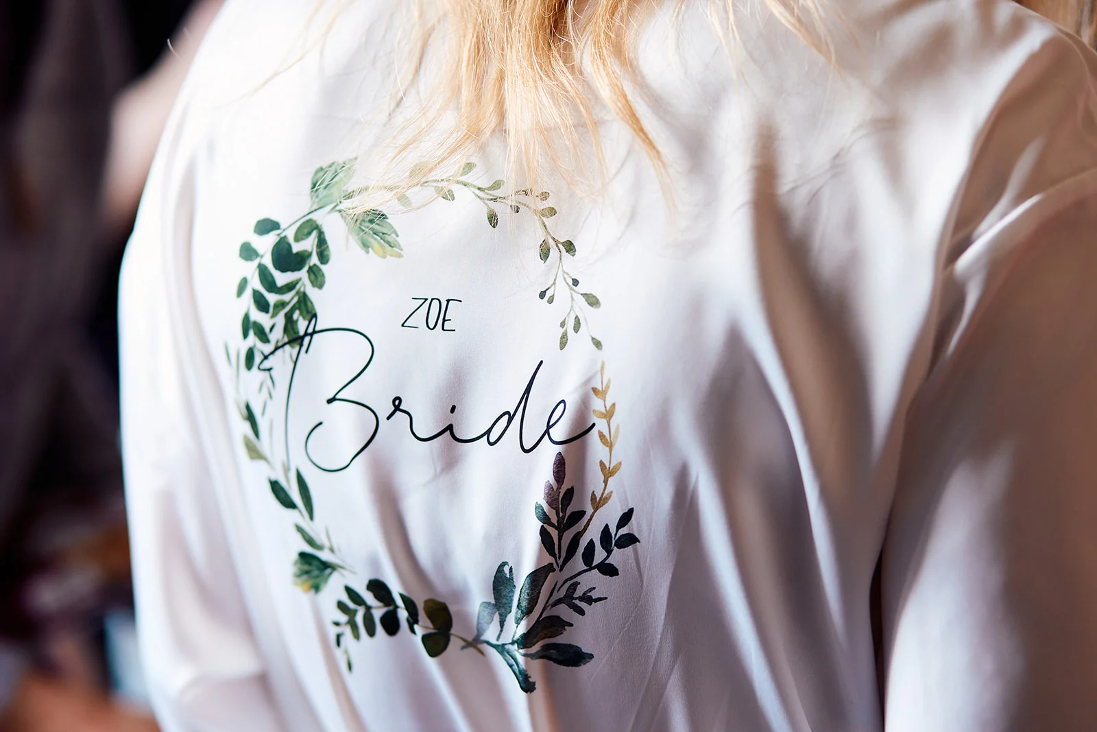 A close-up back view of a bride wearing a personalised white silk robe featuring a green botanical wreath print and the word bride.