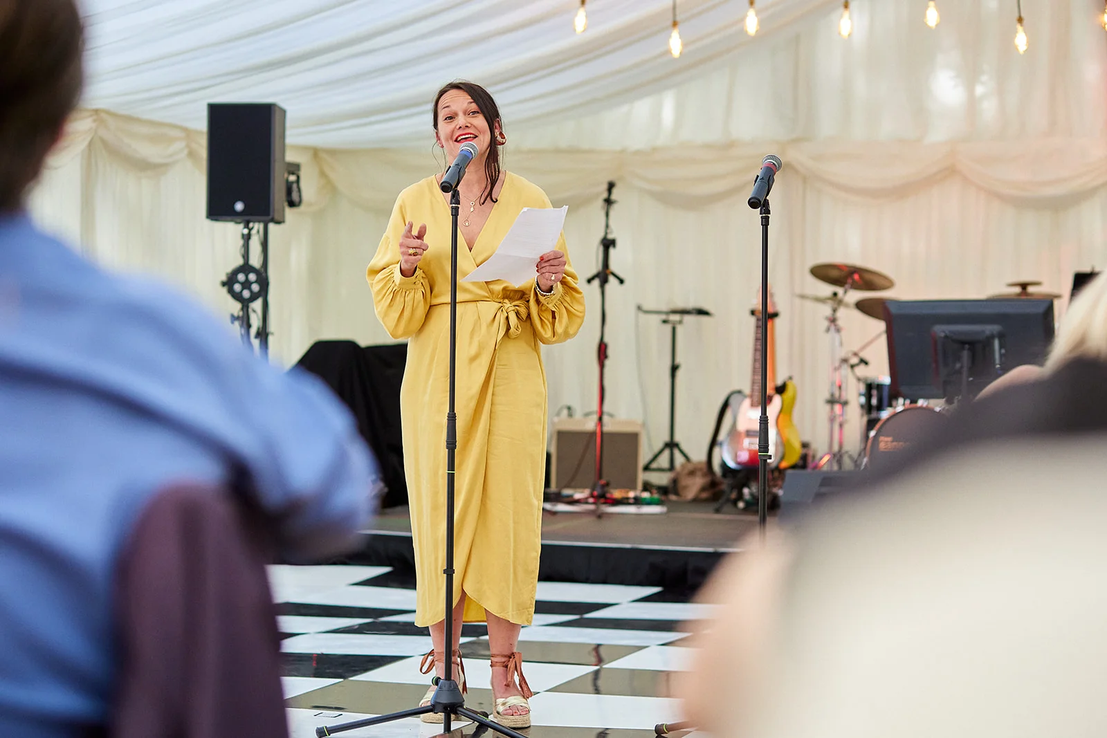 A guest delivers an emotional wedding speech holding notes into a microphone inside a draped marquee with a checkered dance floor.