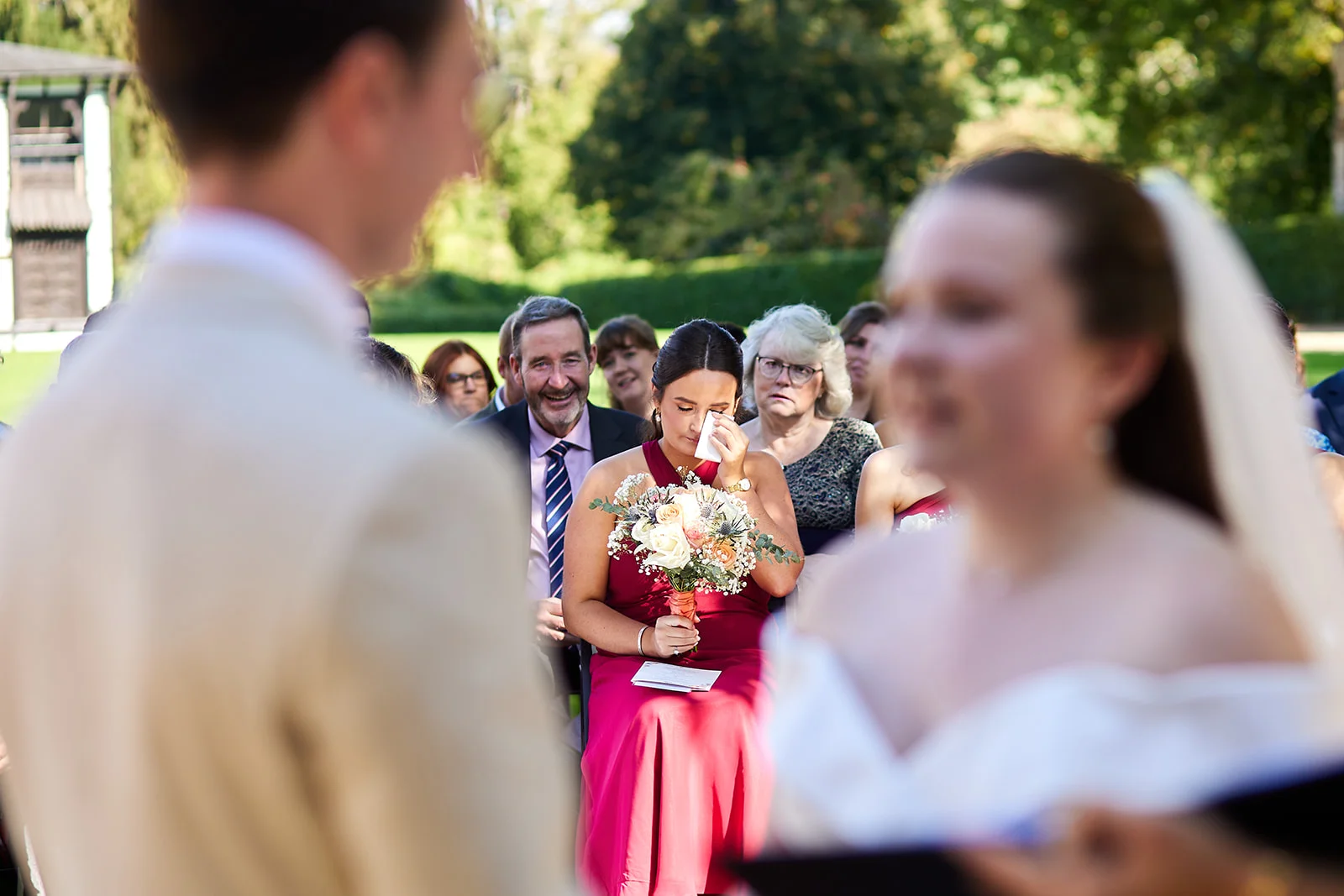 An emotional bridesmaid wipes away a tear with a tissue during an outdoor wedding ceremony while guests look on.