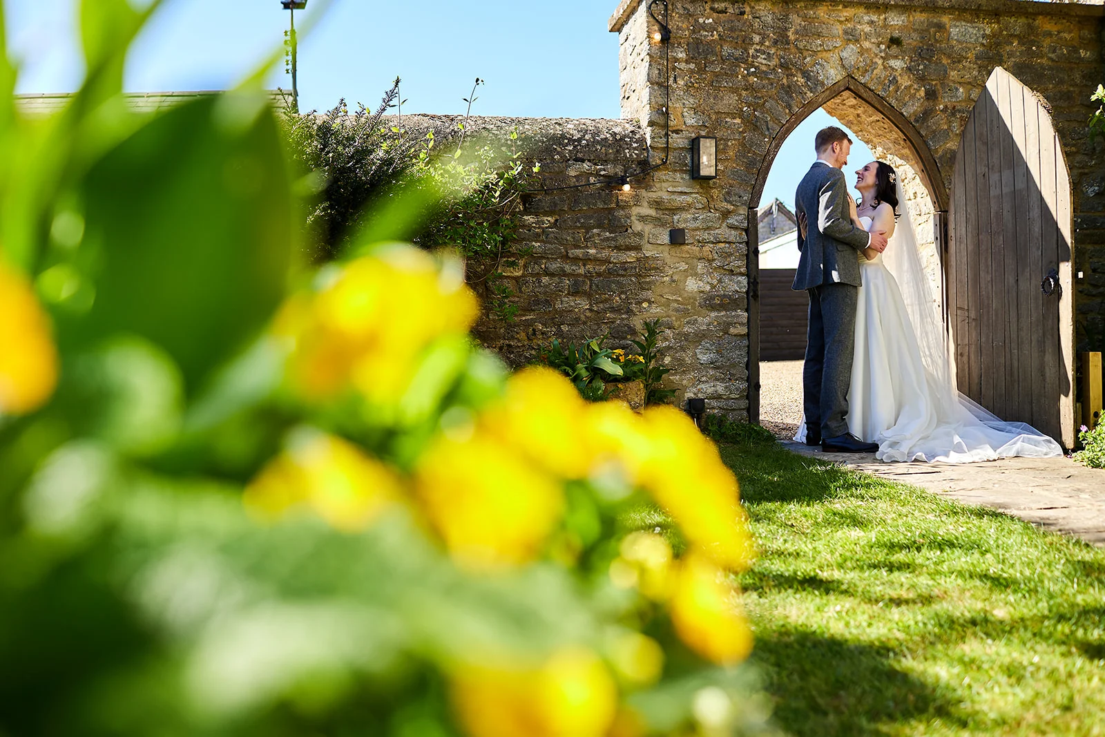 A bride and groom share a joyful moment framed inside a stone archway, viewed past a bright foreground of yellow flowers and green foliage.