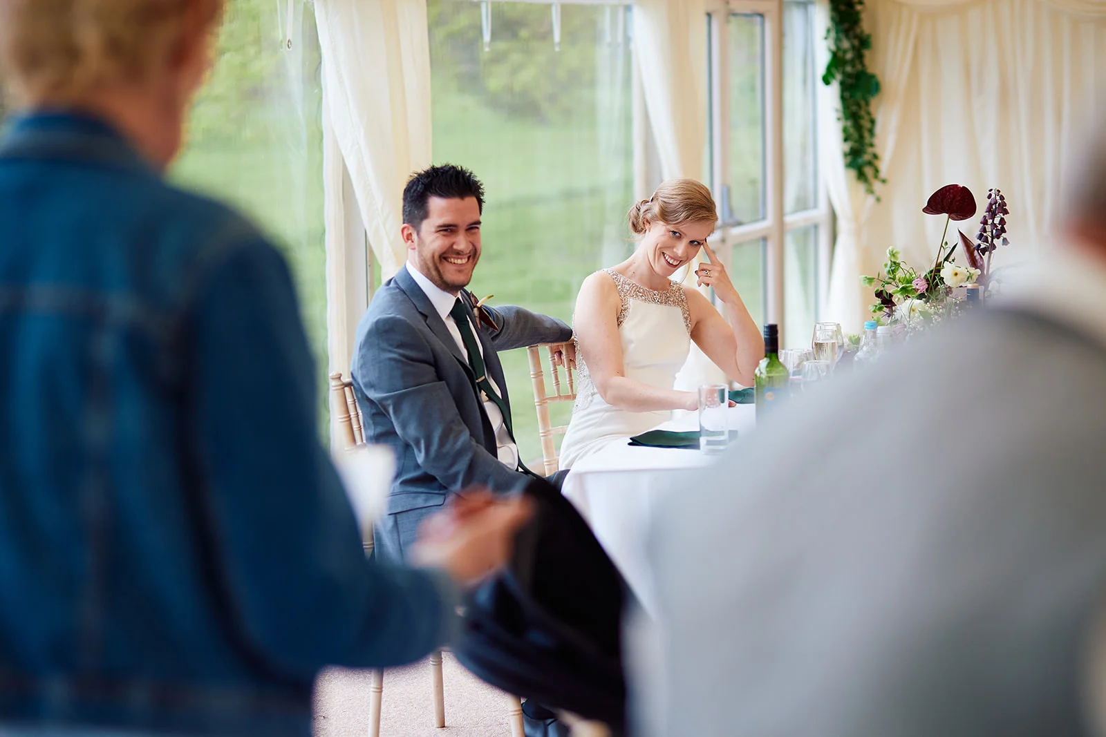The bride and groom laugh together at the top table during the wedding speeches inside a bright marquee venue.