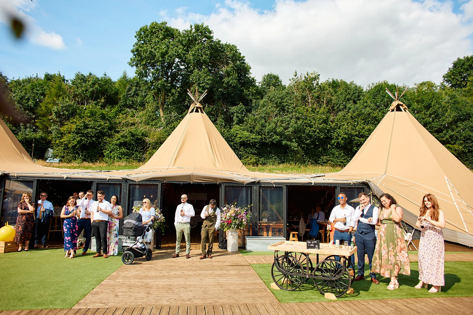 Wedding guests mingle outside large canvas tipi tents under a bright blue summer sky during an outdoor drinks reception.