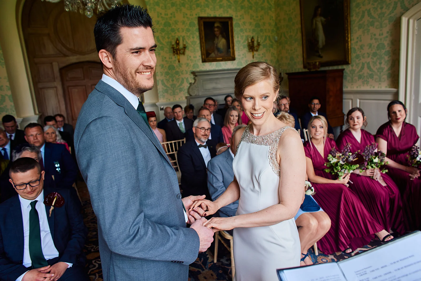 A bride and groom exchange rings during an indoor civil ceremony inside a historic country house, surrounded by seated guests and bridesmaids wearing burgundy dresses.