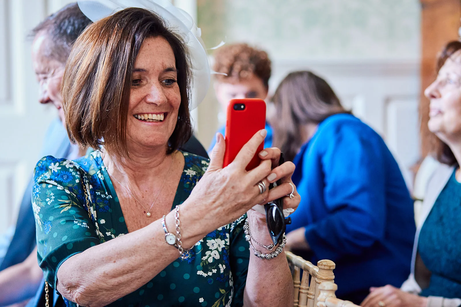 A smiling female wedding guest wearing a floral patterned green dress and a white fascinator takes a photo with her red smartphone during the ceremony.