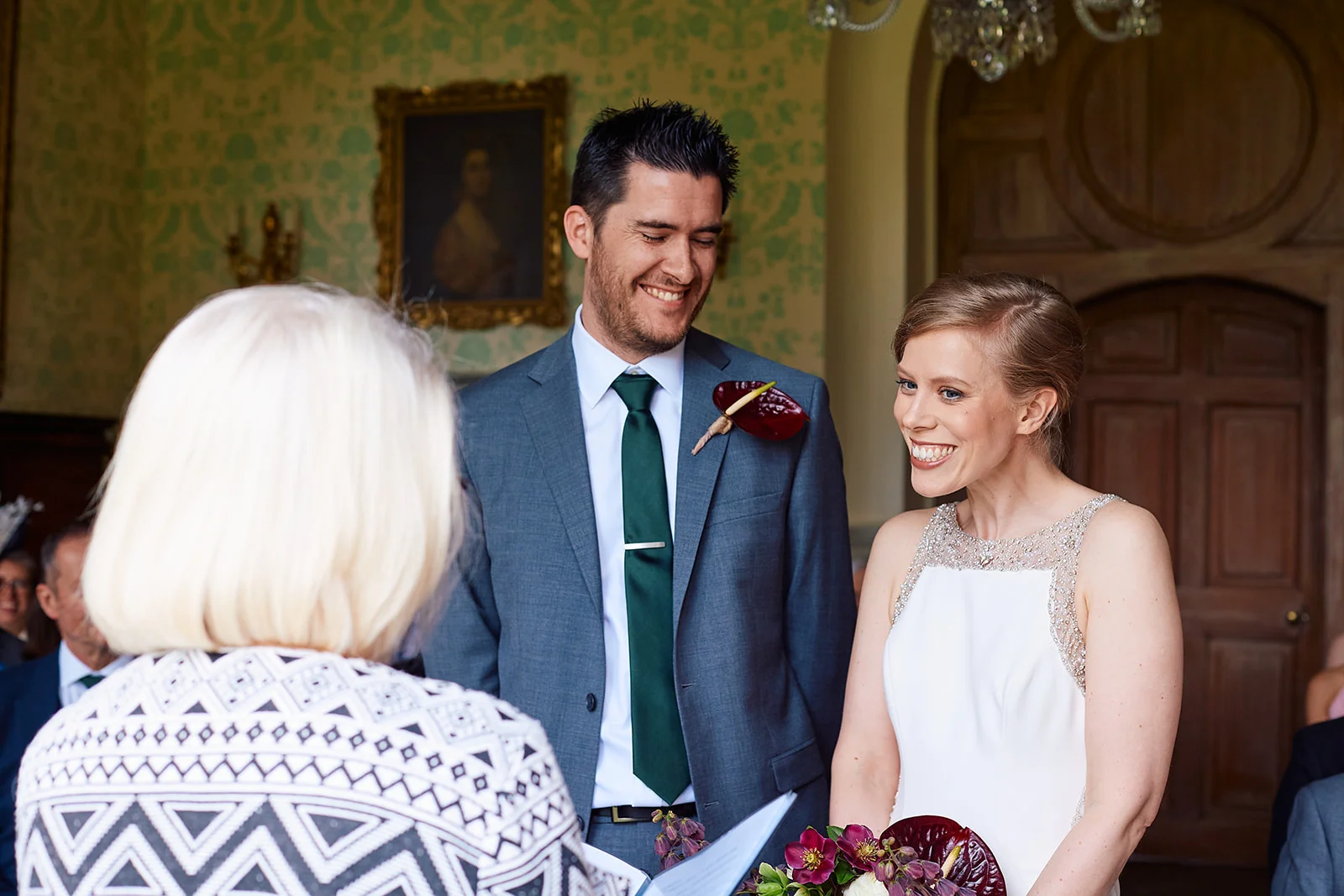 A smiling bride and groom stand together during their indoor civil ceremony, looking joyfully towards the celebrant.