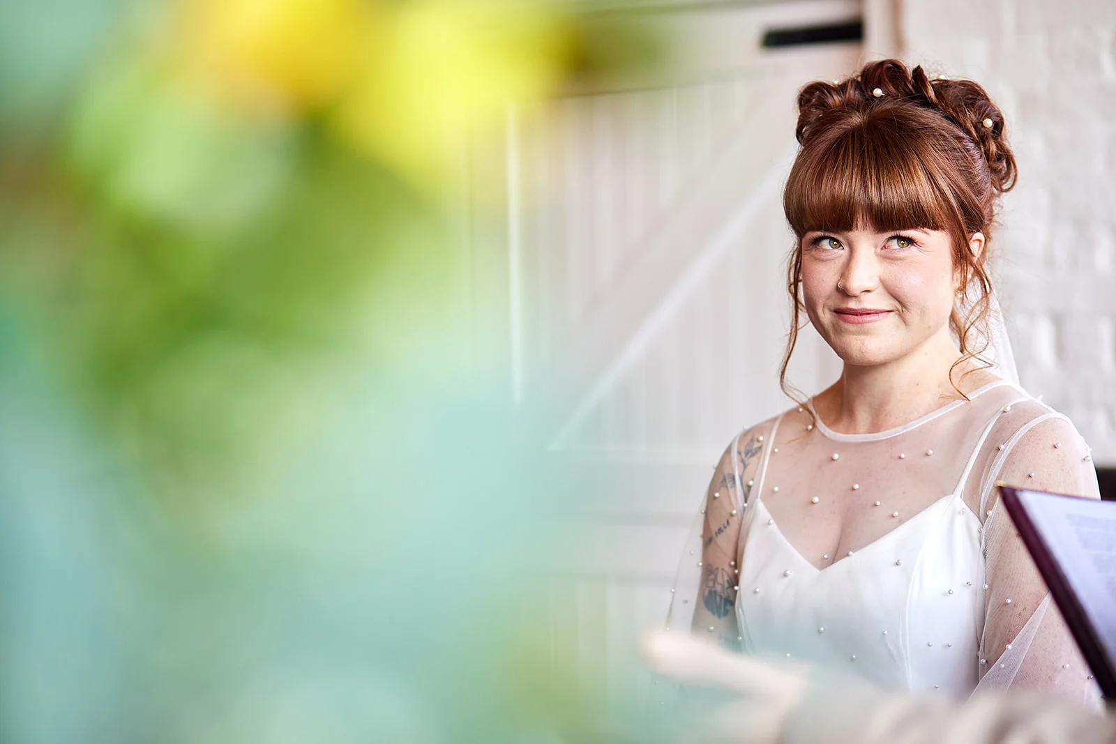 A red-haired bride with an updo and pearl-embellished gown smiles during her wedding ceremony, framed by blurred foreground foliage.