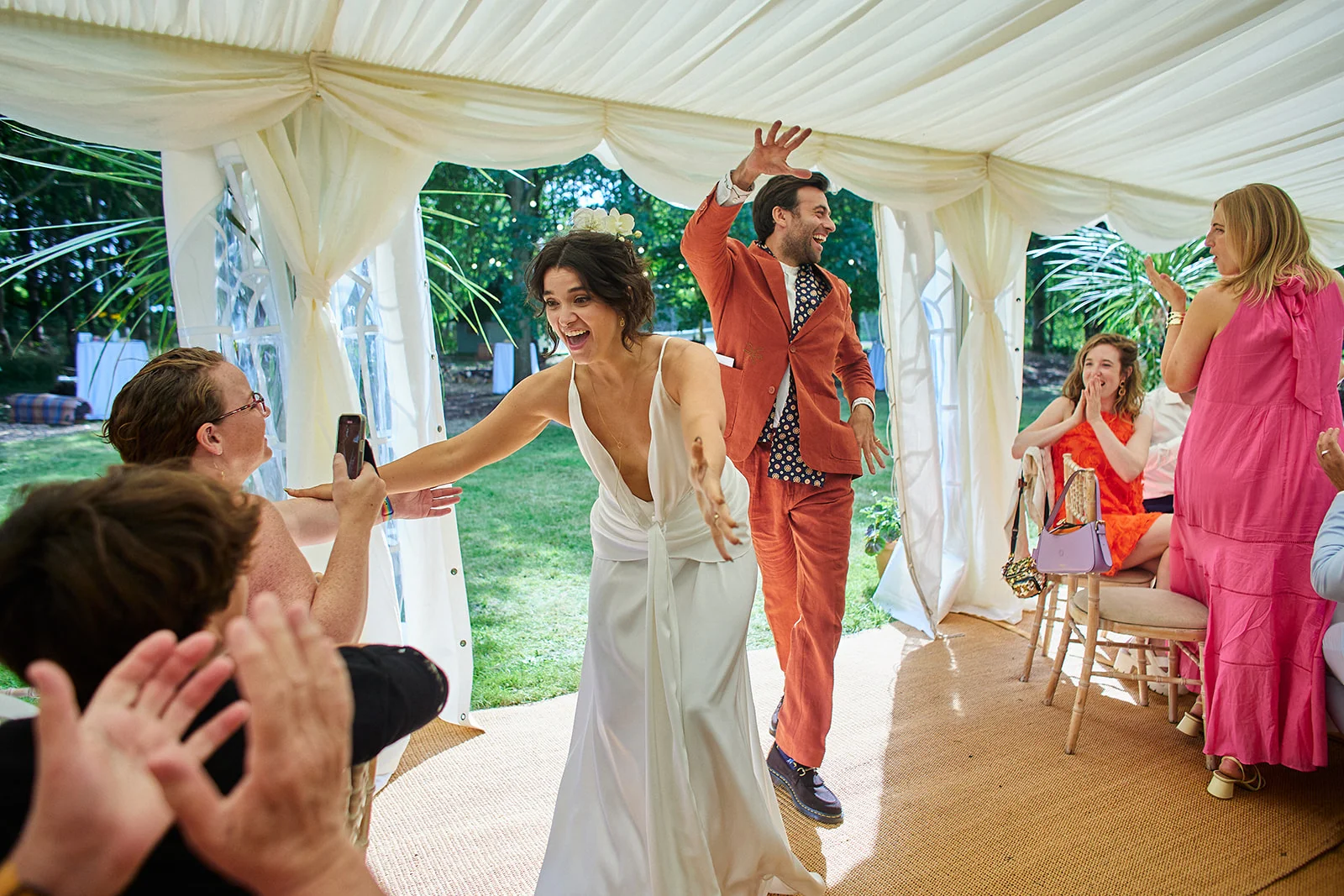 A joyful newlywed couple enter a draped marquee hand in hand, with the laughing bride wearing a plunging slip dress and the groom in a rust suit as guests cheer and applaud.
