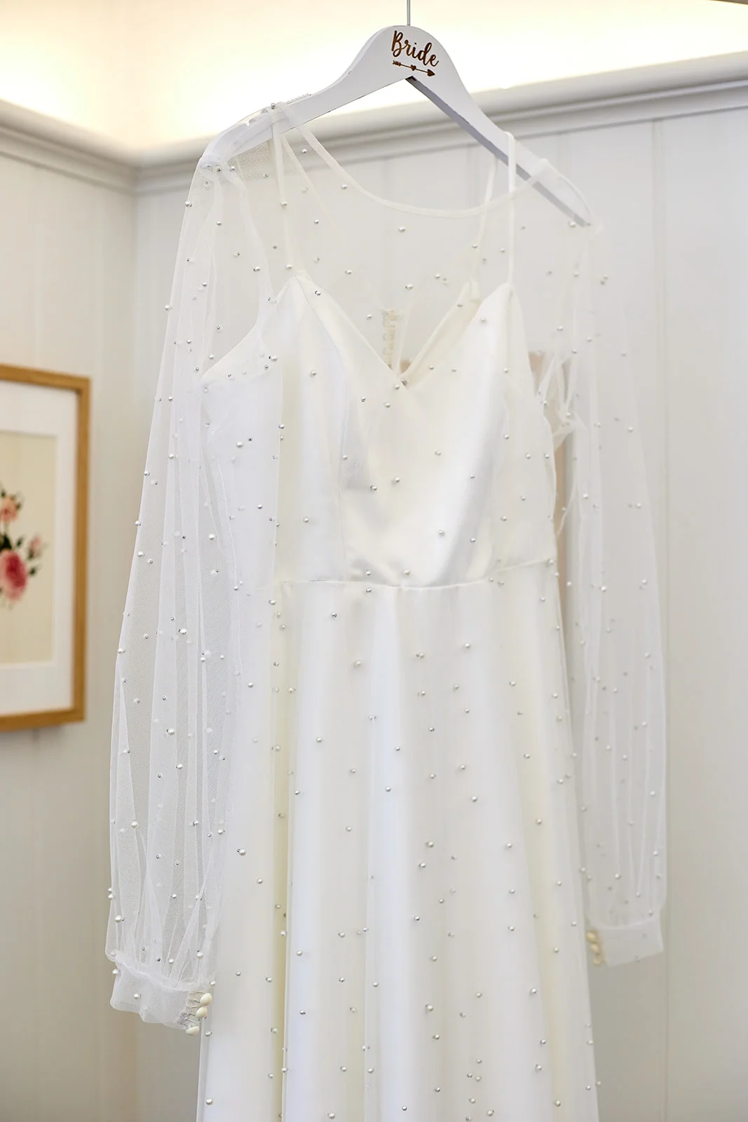 A stunning pearl-encrusted long-sleeve wedding gown with a delicate sheer overlay hangs delicately on a personalised hanger during bridal preparations.
