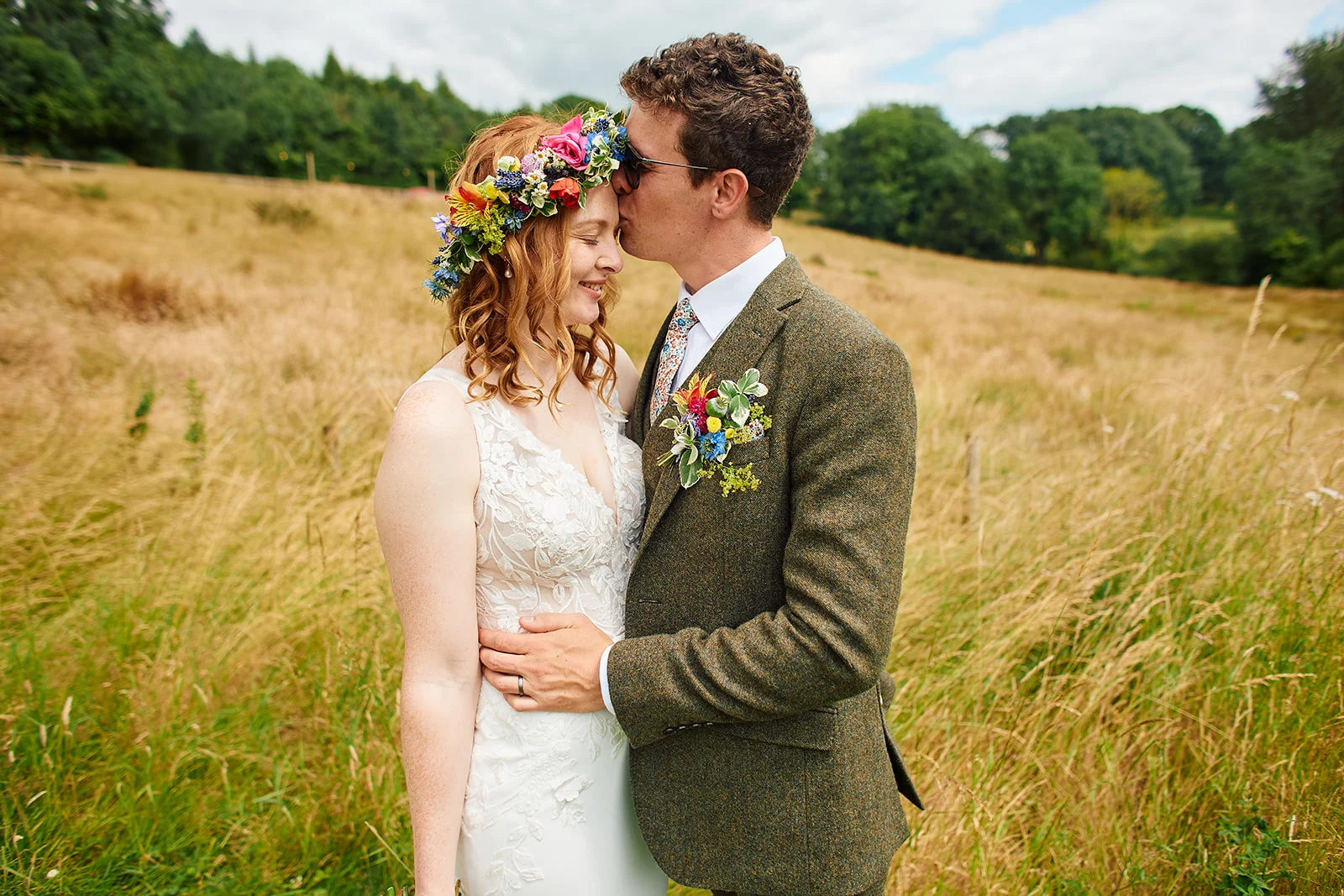 A groom wearing a tweed suit and sunglasses gently kisses his bride on the forehead as they stand in a scenic wildflower field.
