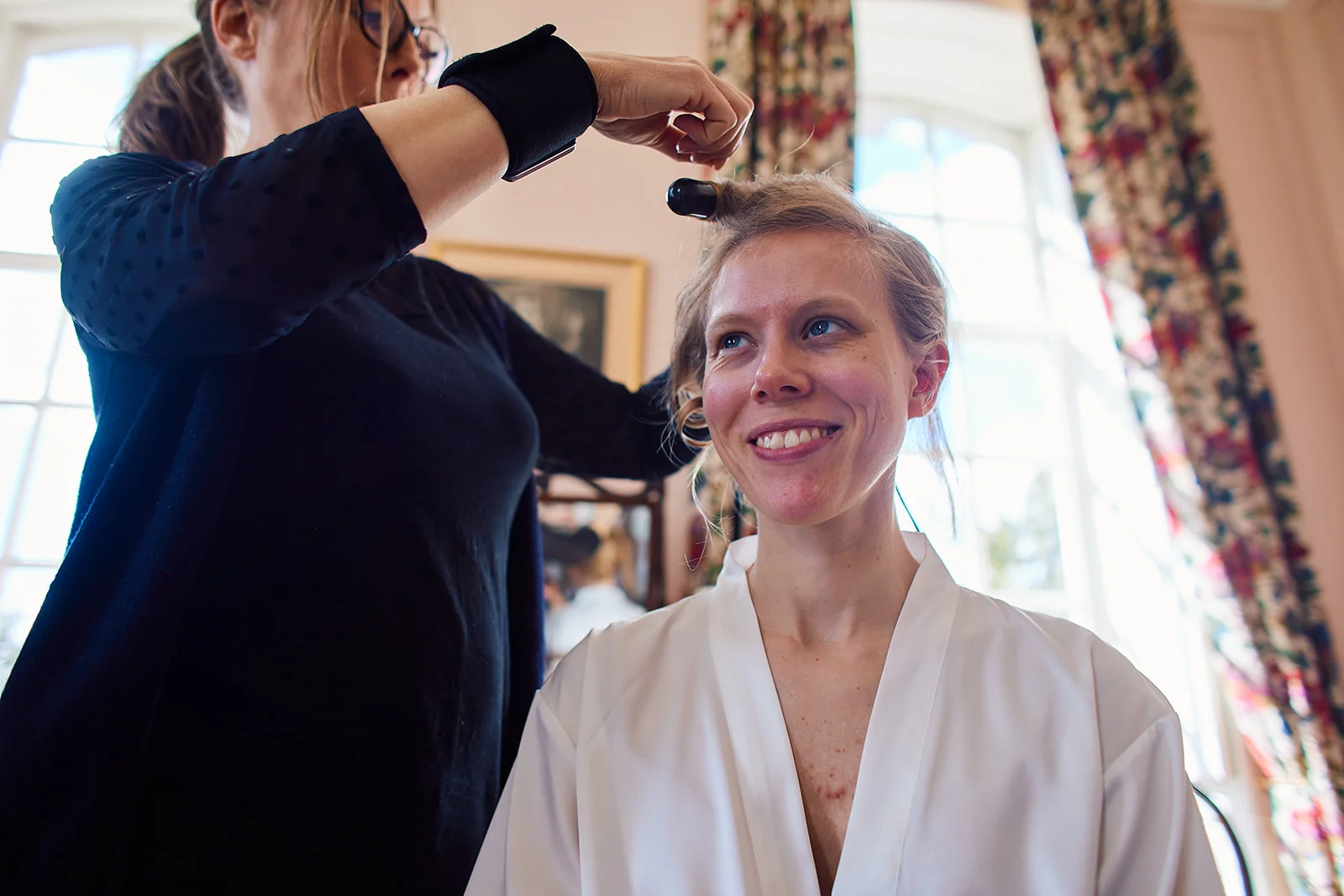 A smiling bride wearing a white robe has her hair styled by a professional against a bright window backdrop during wedding morning preparations.