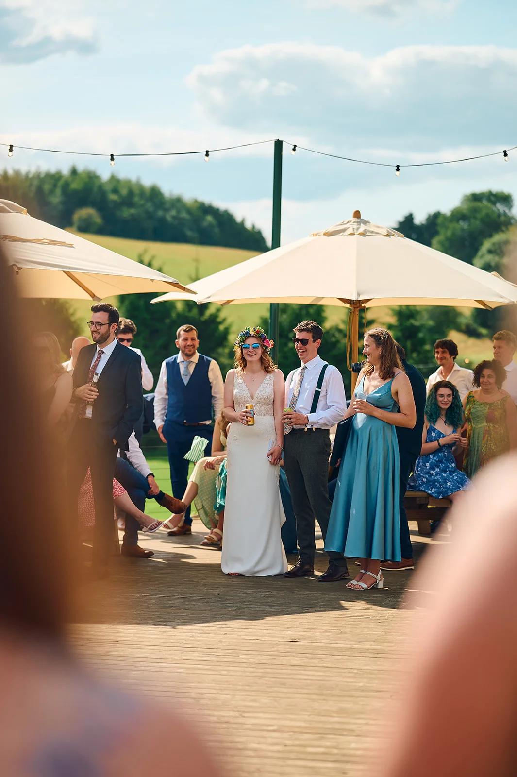 A bride in a white gown and flower crown laughs with wedding guests during an outdoor drinks reception on a sunny day.