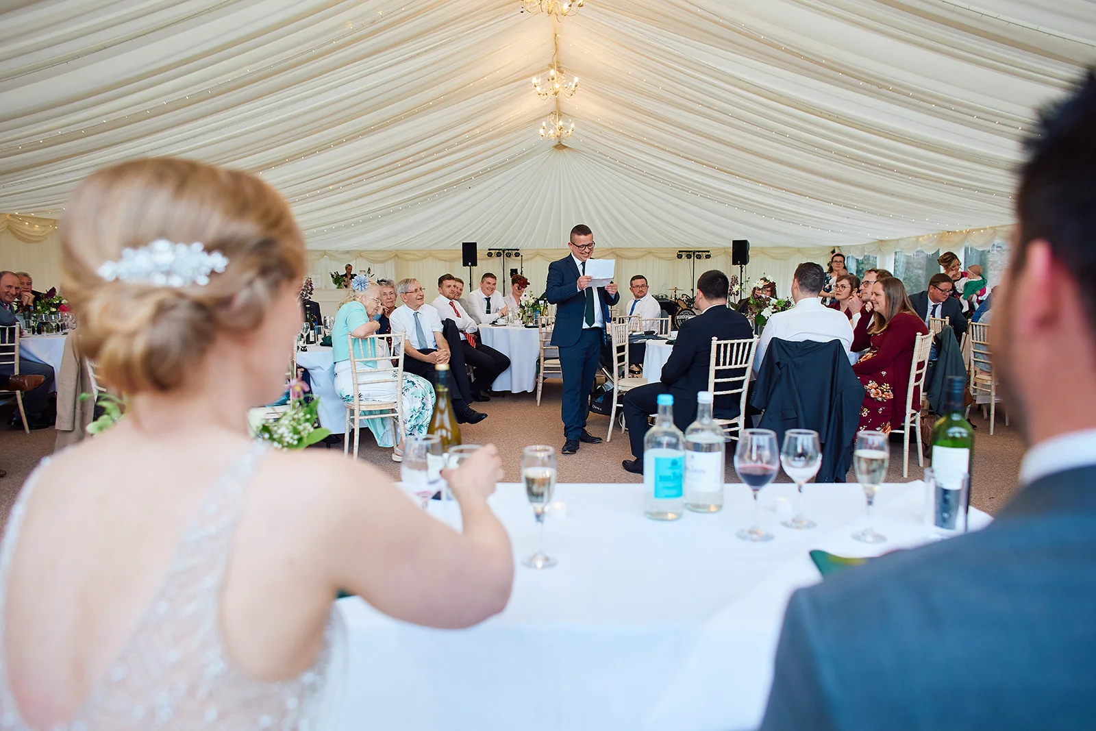 A groom delivers a speech while holding notes inside a large draped marquee wedding reception, viewed over the shoulders of the seated bride and groom.