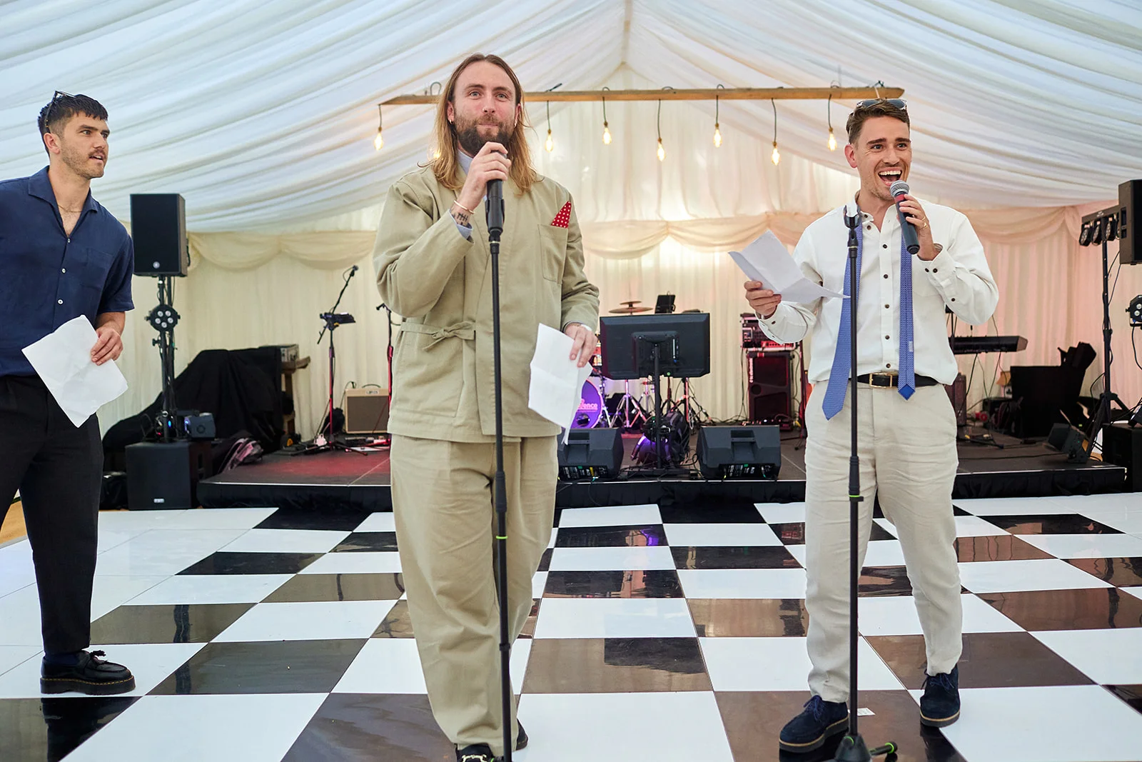 Three men stand on a black and white checkered dance floor inside a draped marquee, holding microphones and speaking during wedding speeches.