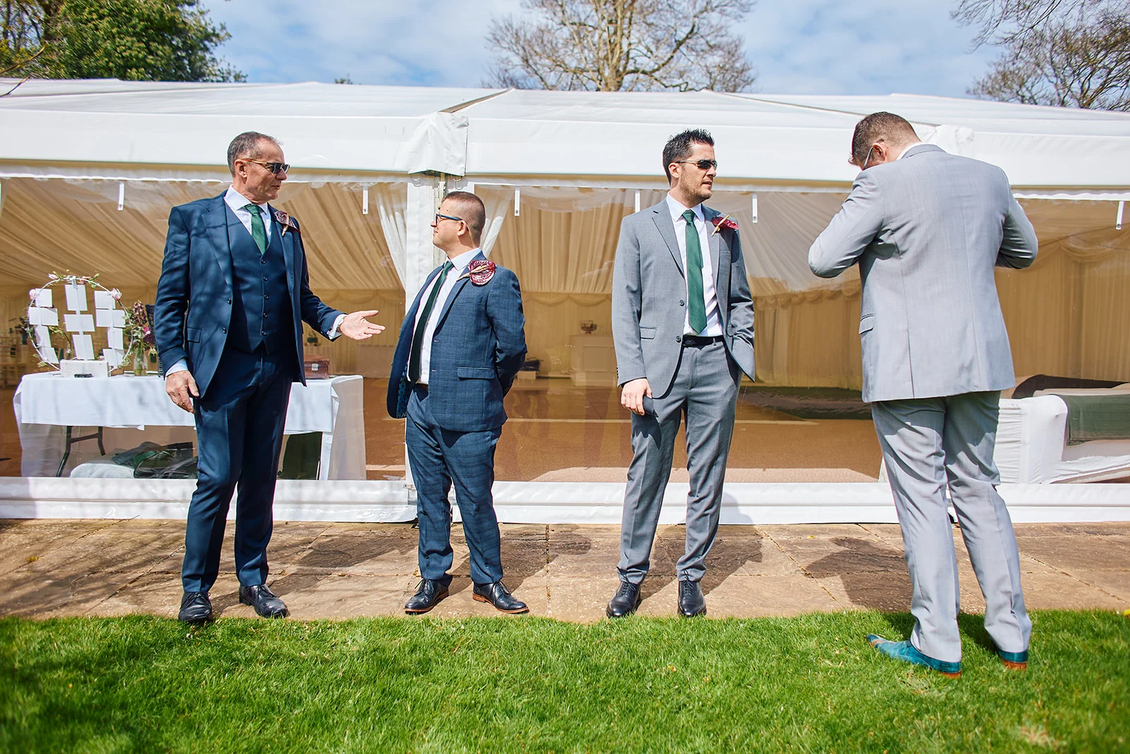 A group of smartly dressed groomsmen in suits stand outside a wedding marquee on a bright sunny day.