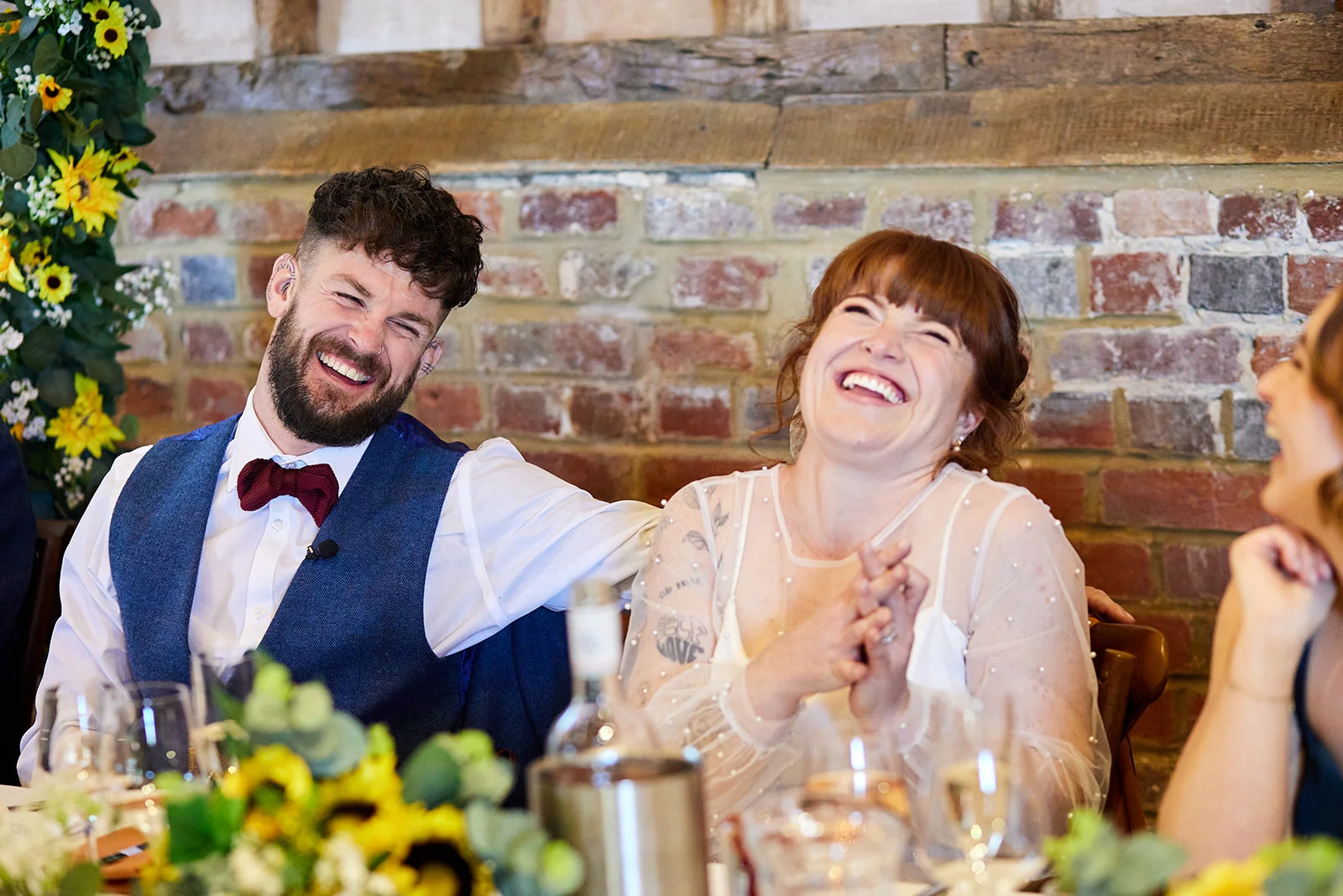 A bride and groom share a joyful laugh while seated at the top table during wedding speeches in a rustic barn venue.