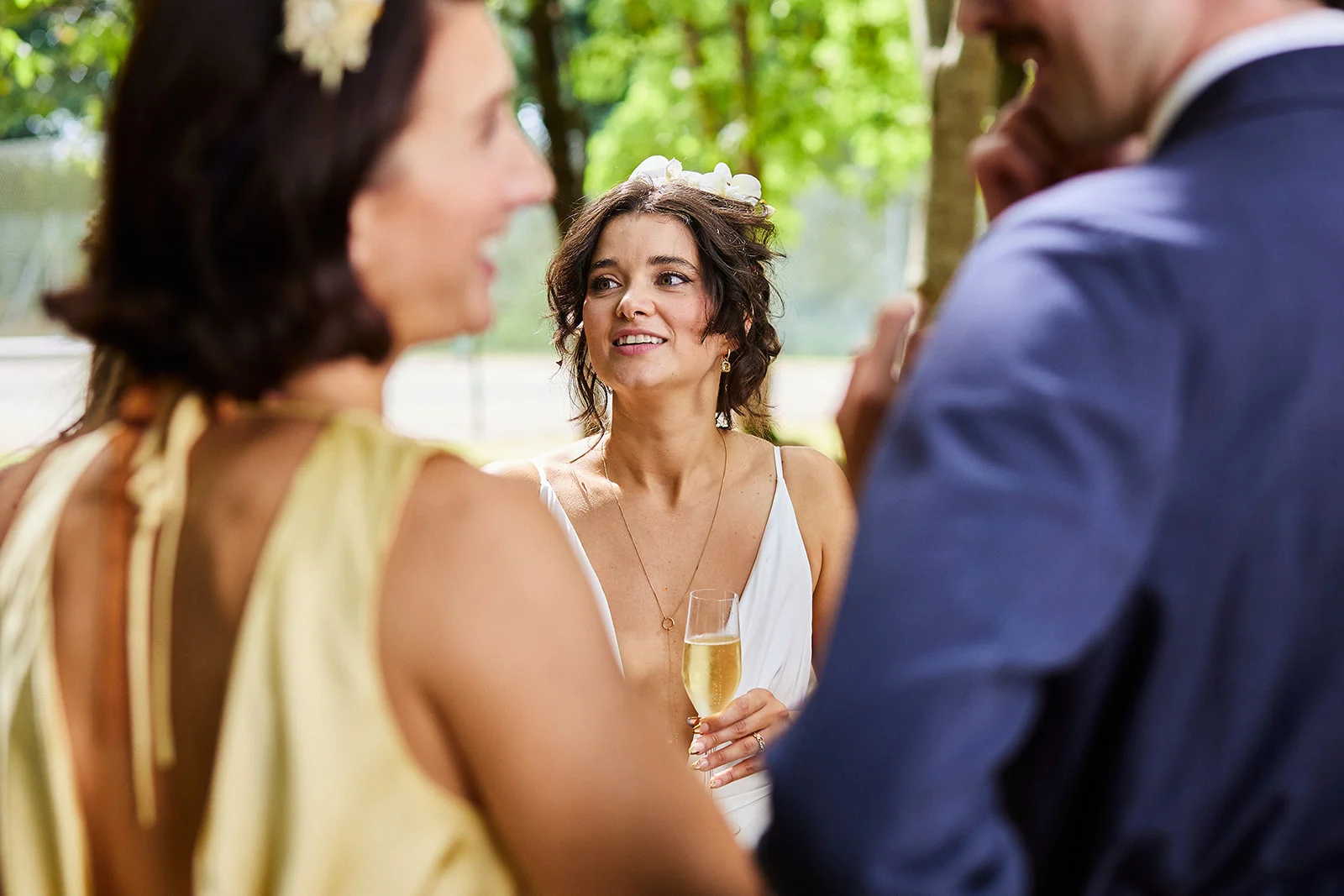 A guest holding a glass of champagne smiles during an outdoor wedding drinks reception, surrounded by chatting attendees.