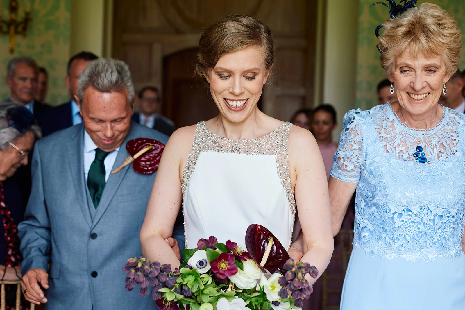 A smiling bride walks down the aisle holding a vibrant bouquet, accompanied by her parents in a bright indoor ceremony setting.