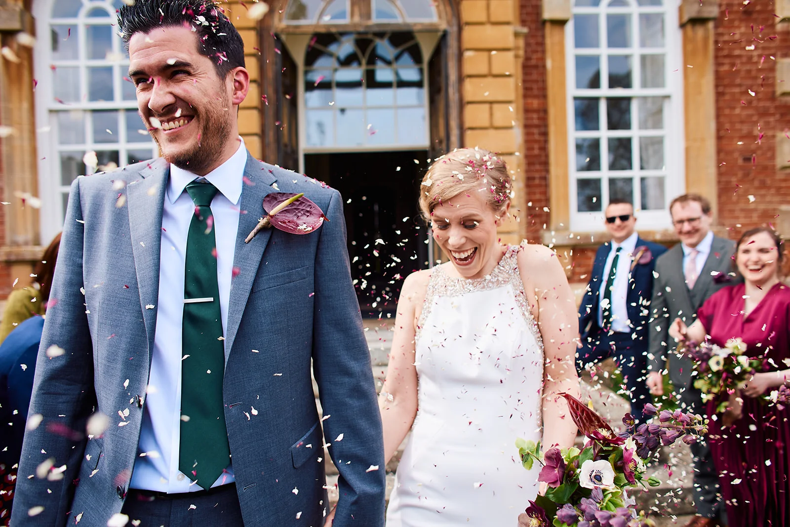 A joyful newlywed couple walk hand in hand through a shower of confetti outside a grand brick country house.