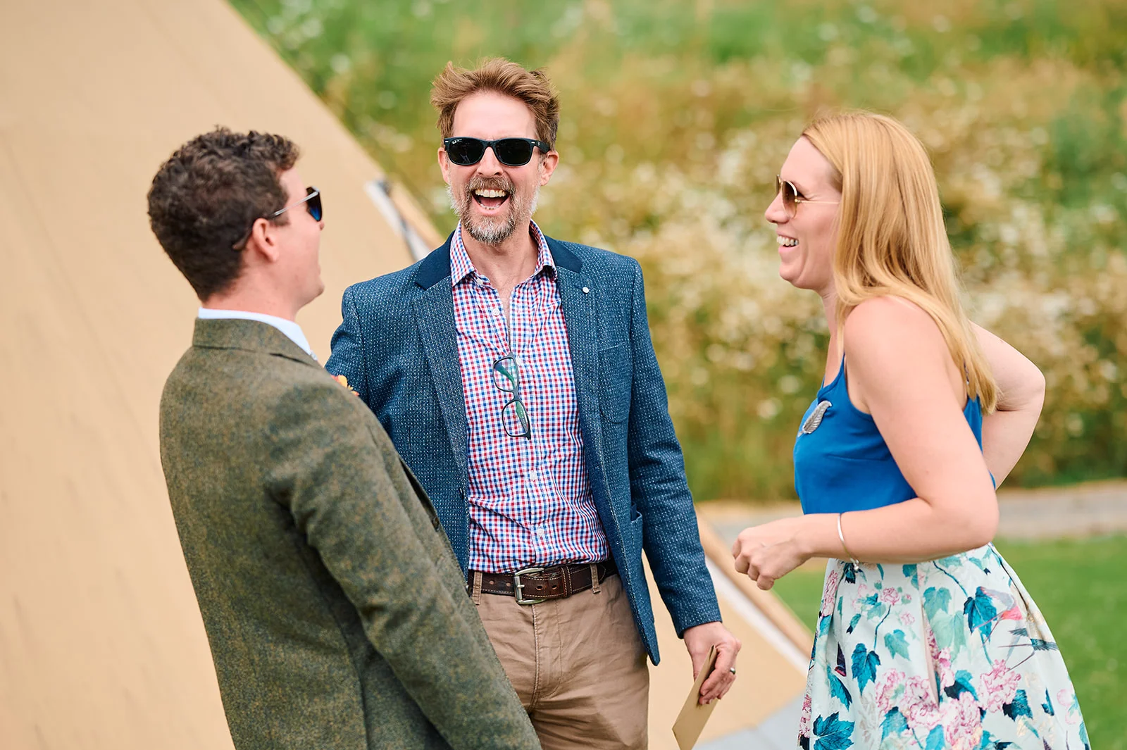 Three wedding guests share a joyful conversation outside a tipi venue on a bright summer afternoon.
